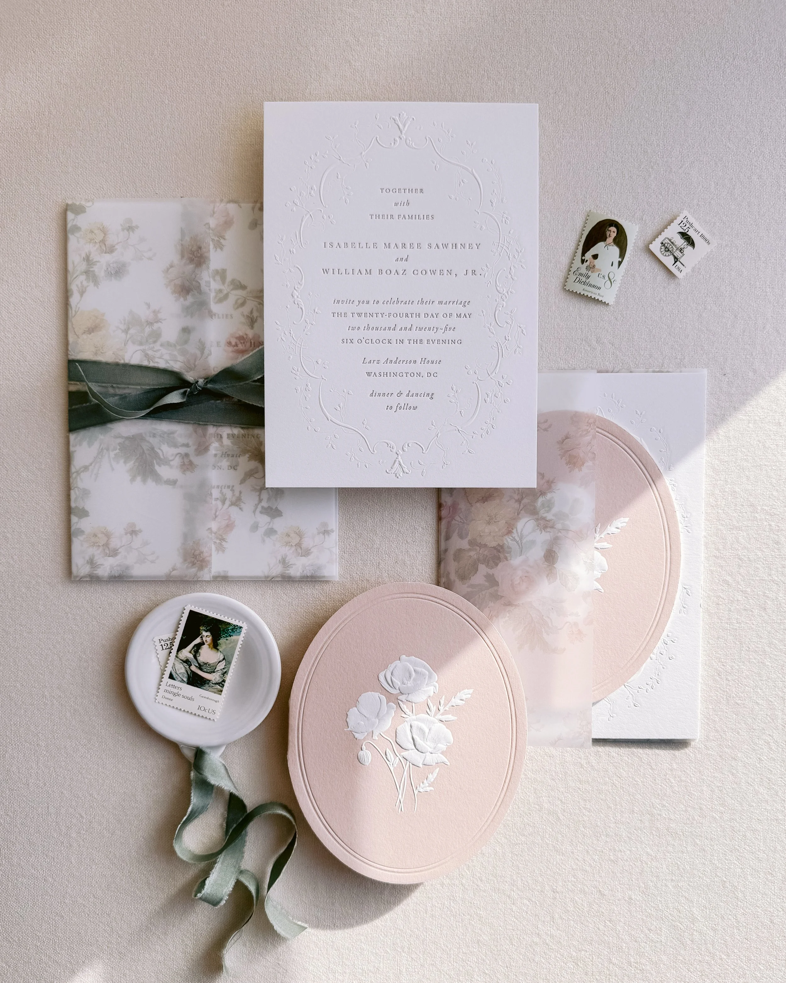 Wedding invitation with floral details, wrapped in floral paper and ribbon, pink floral box, mailing stamps, and a wax seal with a ribbon, all arranged on a light-colored surface.