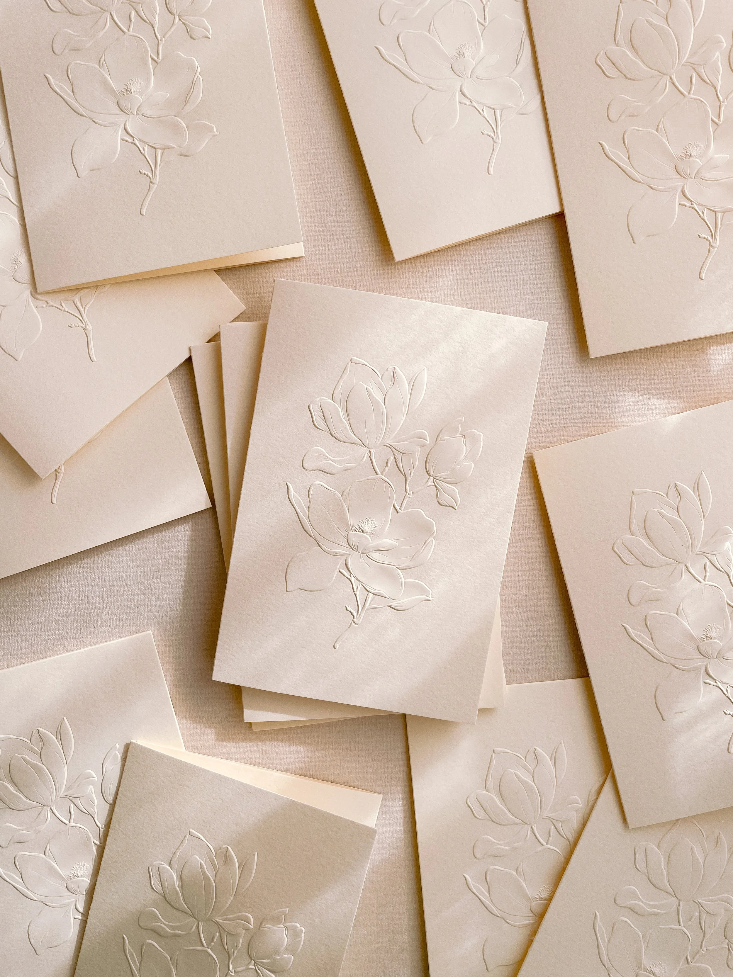 Multiple cream-colored cards with embossed magnolia flower designs, some stacked and some spread out on a light surface.
