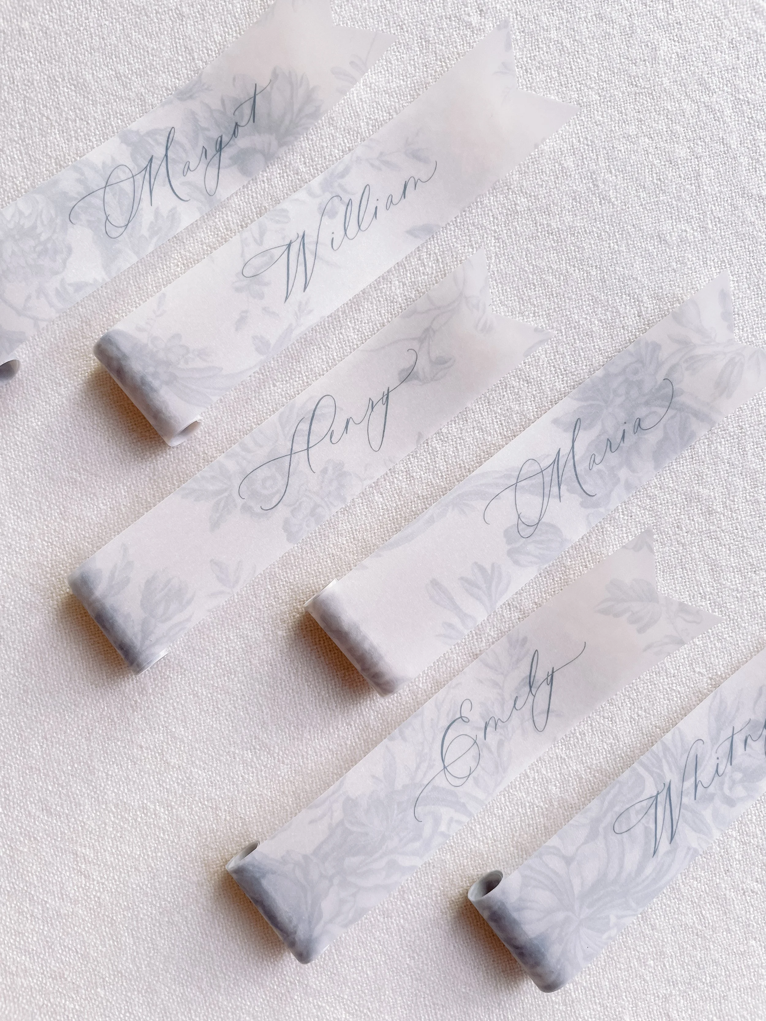 Seven white paper name tags with elegant gray cursive writing on a white textured surface. The tags read 'Margo,' 'William,' 'Amy,' 'Maria,' 'Sam,' and 'Whitney.'
