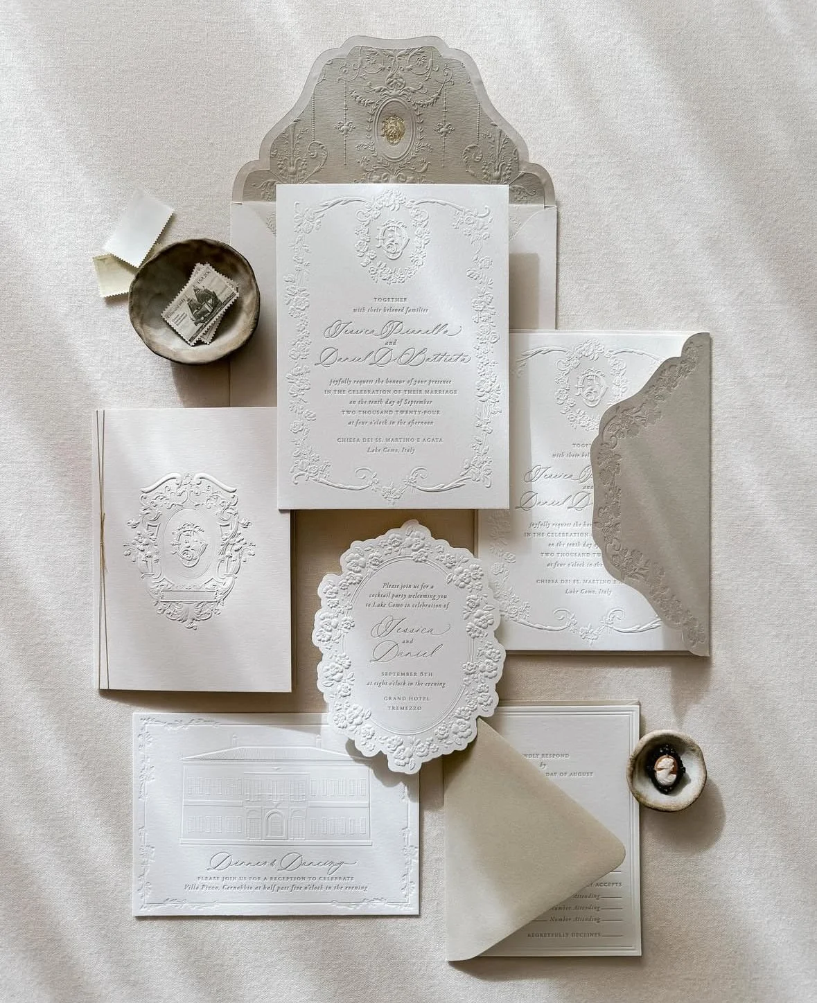 Elegant white wedding invitations and RSVP cards with embossed details, accompanied by a small dish with stamps and a shell-shaped bowl on a white surface.