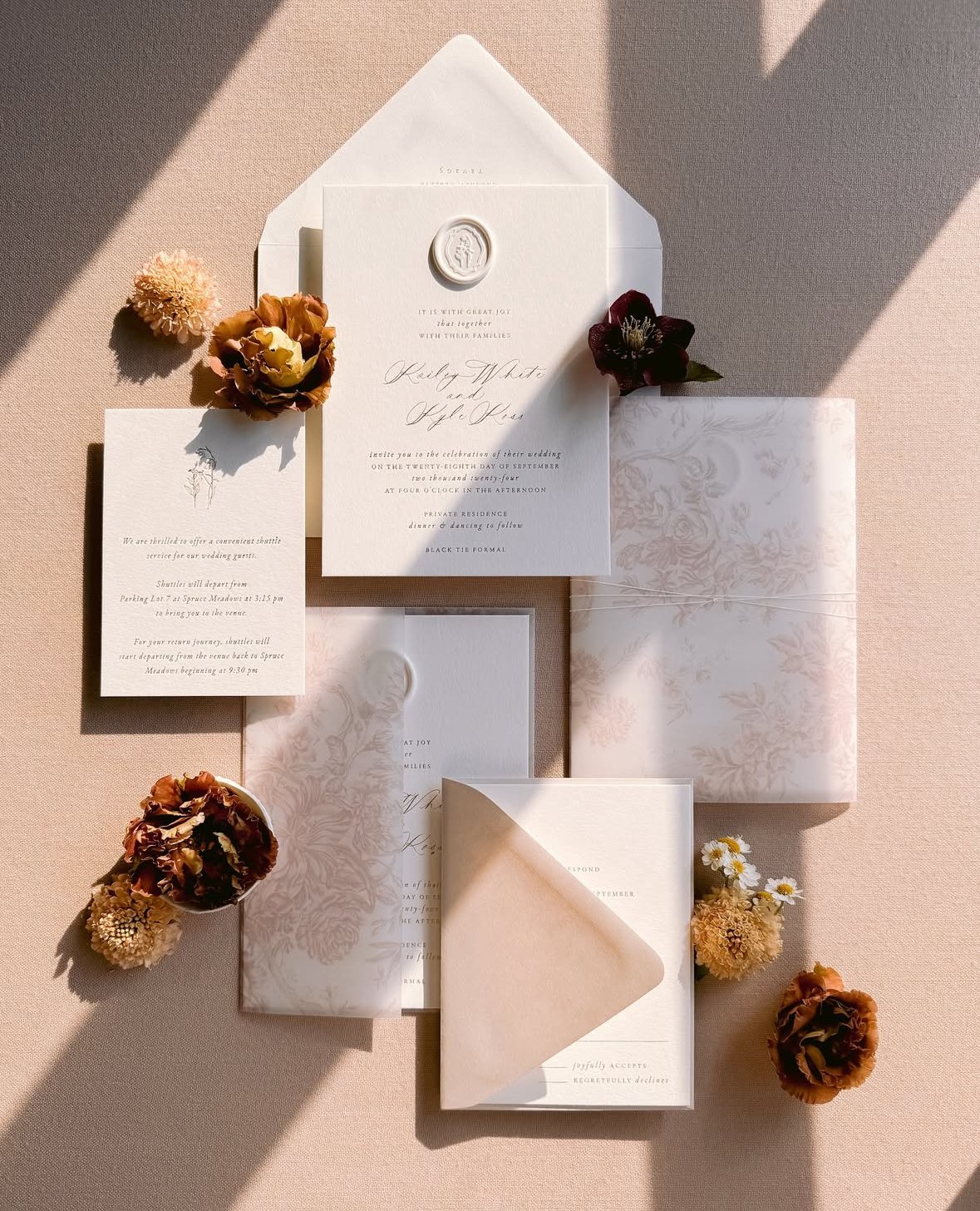 Wedding invitation suite with floral accents and dried flowers on a pink surface, including an invitation, RSVP card, envelopes, and decorative paper.