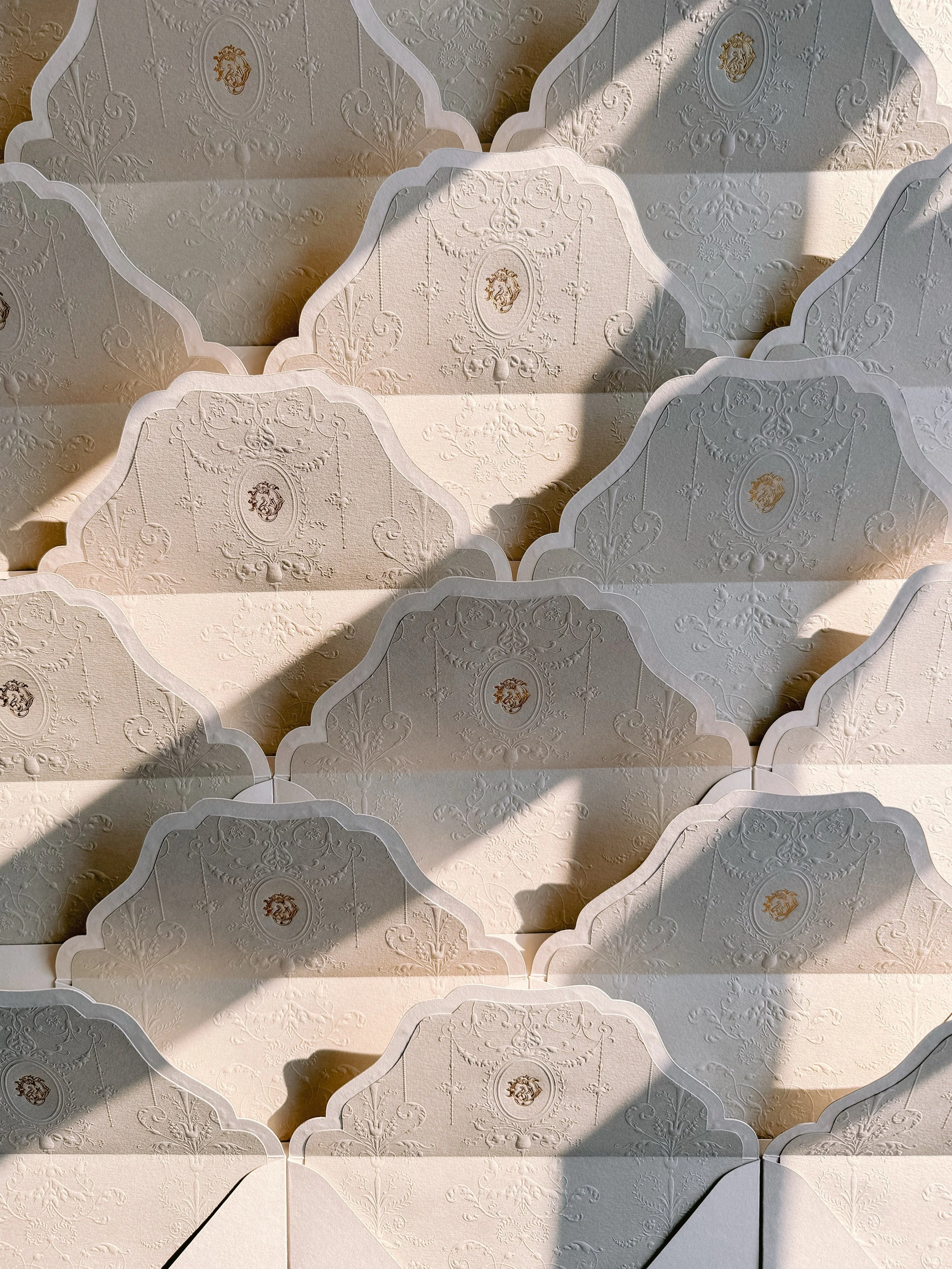 A pattern of white paper or cardboard with embossed ornate designs and a small crest or emblem, arranged in a repeating pattern, with shadows cast over some of the pieces.