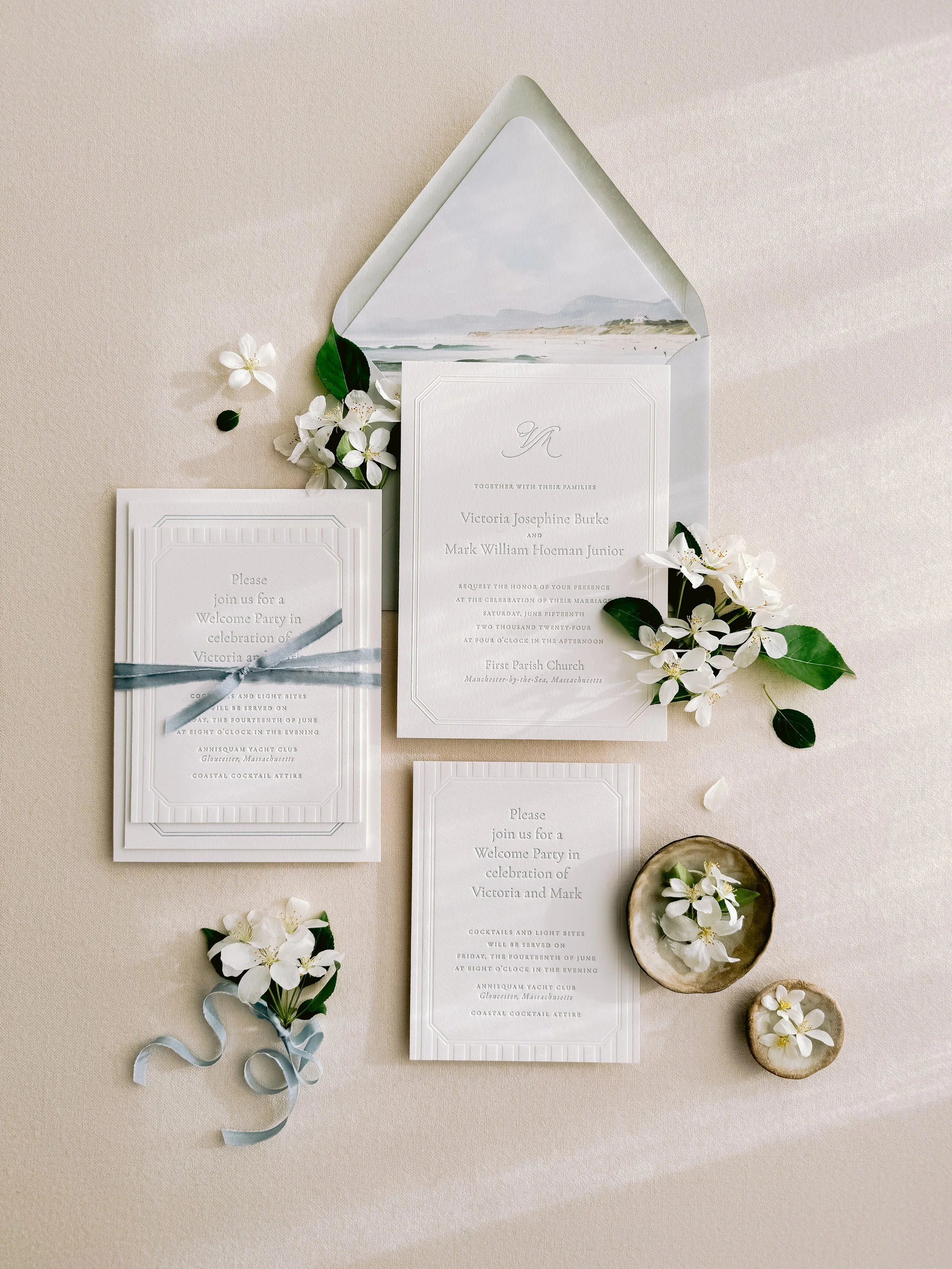 Elegant wedding invitation set with floral accents, including RSVP card, main invitation, and welcome party card, all surrounded by white flowers and greenery on a light background.