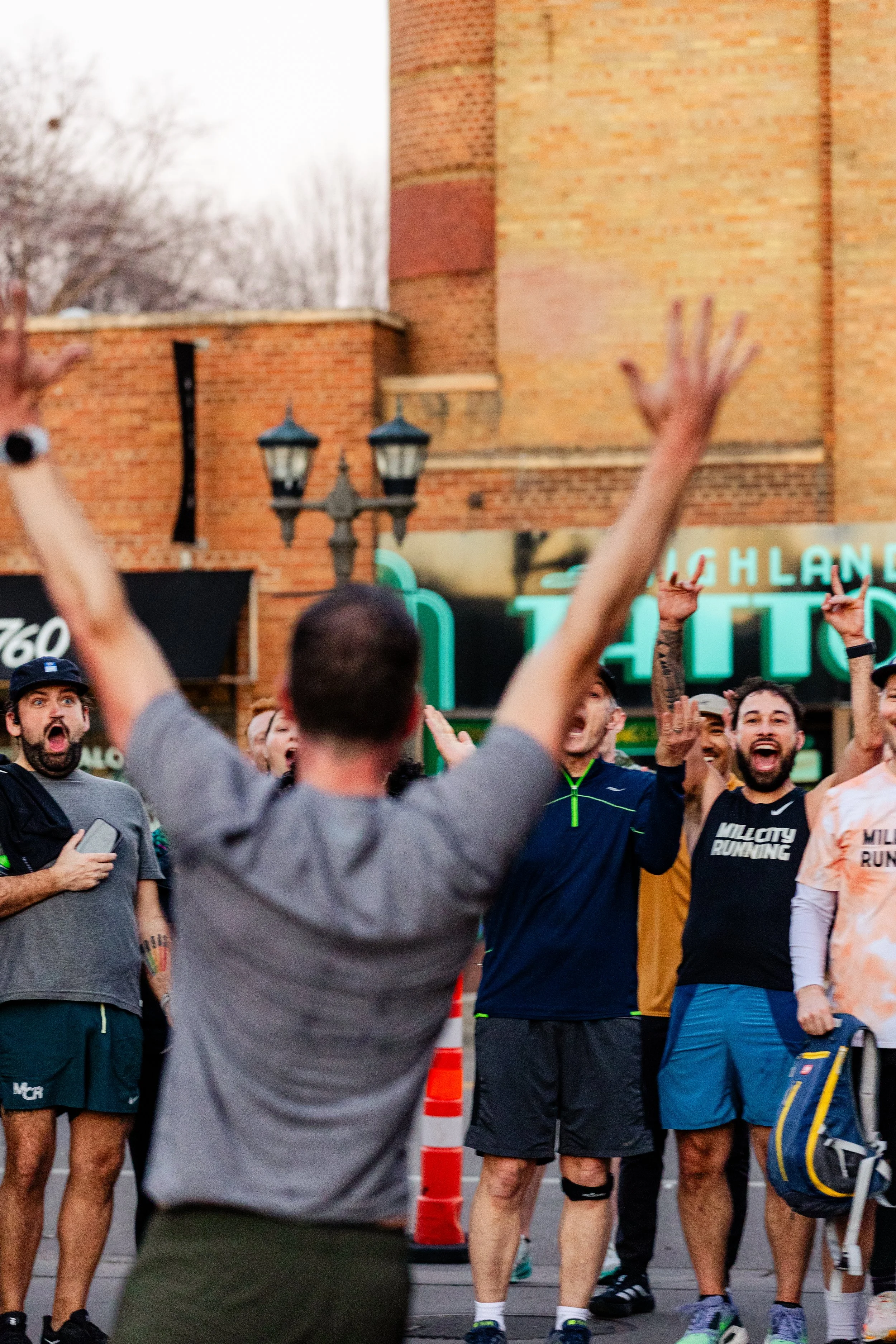A group of people gathered outdoors, some wearing athletic clothing, celebrating or cheering. The person in the foreground has their arms raised. The background shows a brick building and neon signs.
