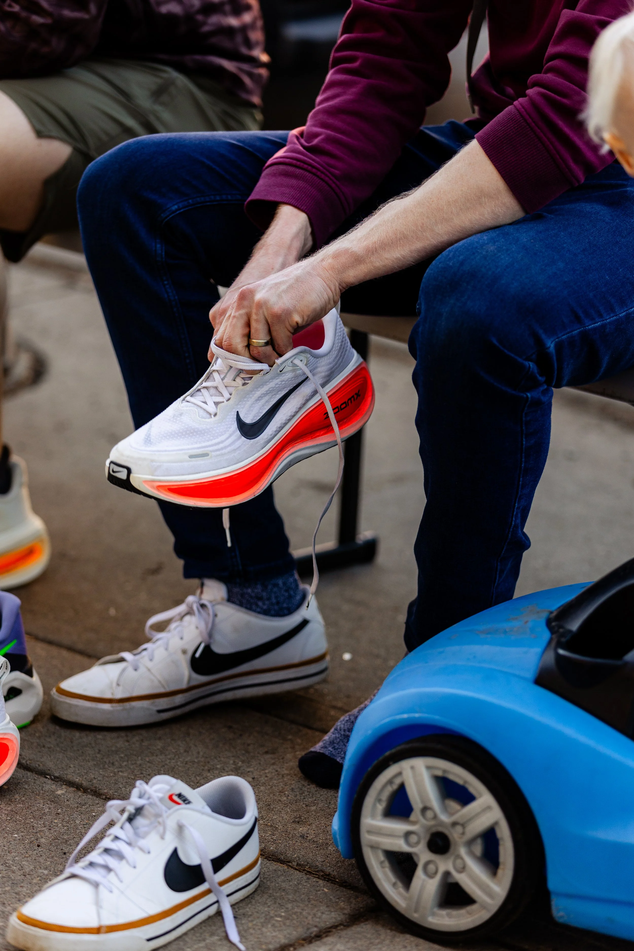 Person sitting on a chair tying their white Nike sneakers with black swoosh logo, orange air cushion sole, wearing blue jeans and a burgundy sweatshirt. There are additional sneakers on the ground and a small blue toy car nearby.