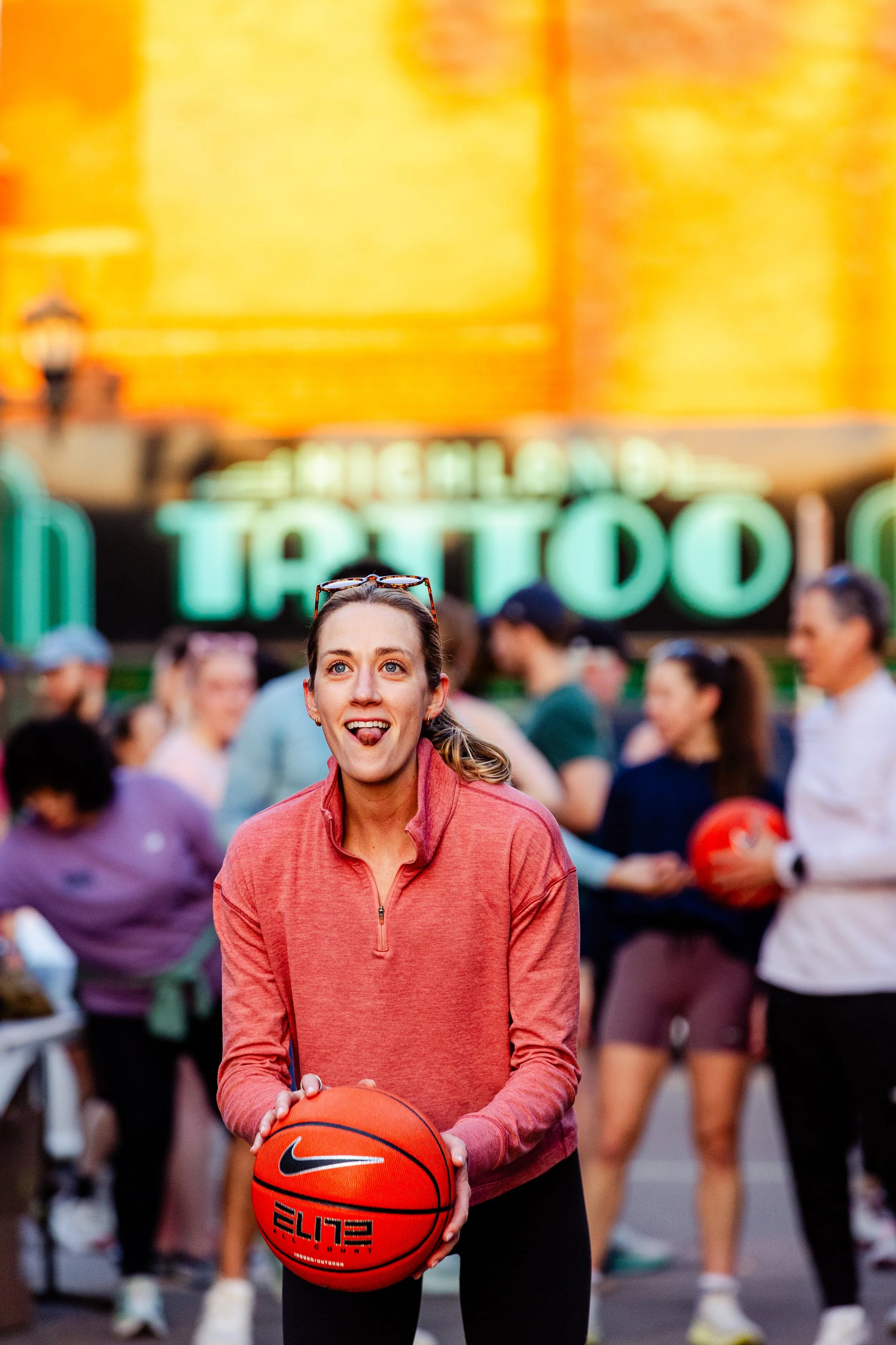 A woman holding a Nike basketball at an outdoor event with a crowd and a neon sign in the background.
