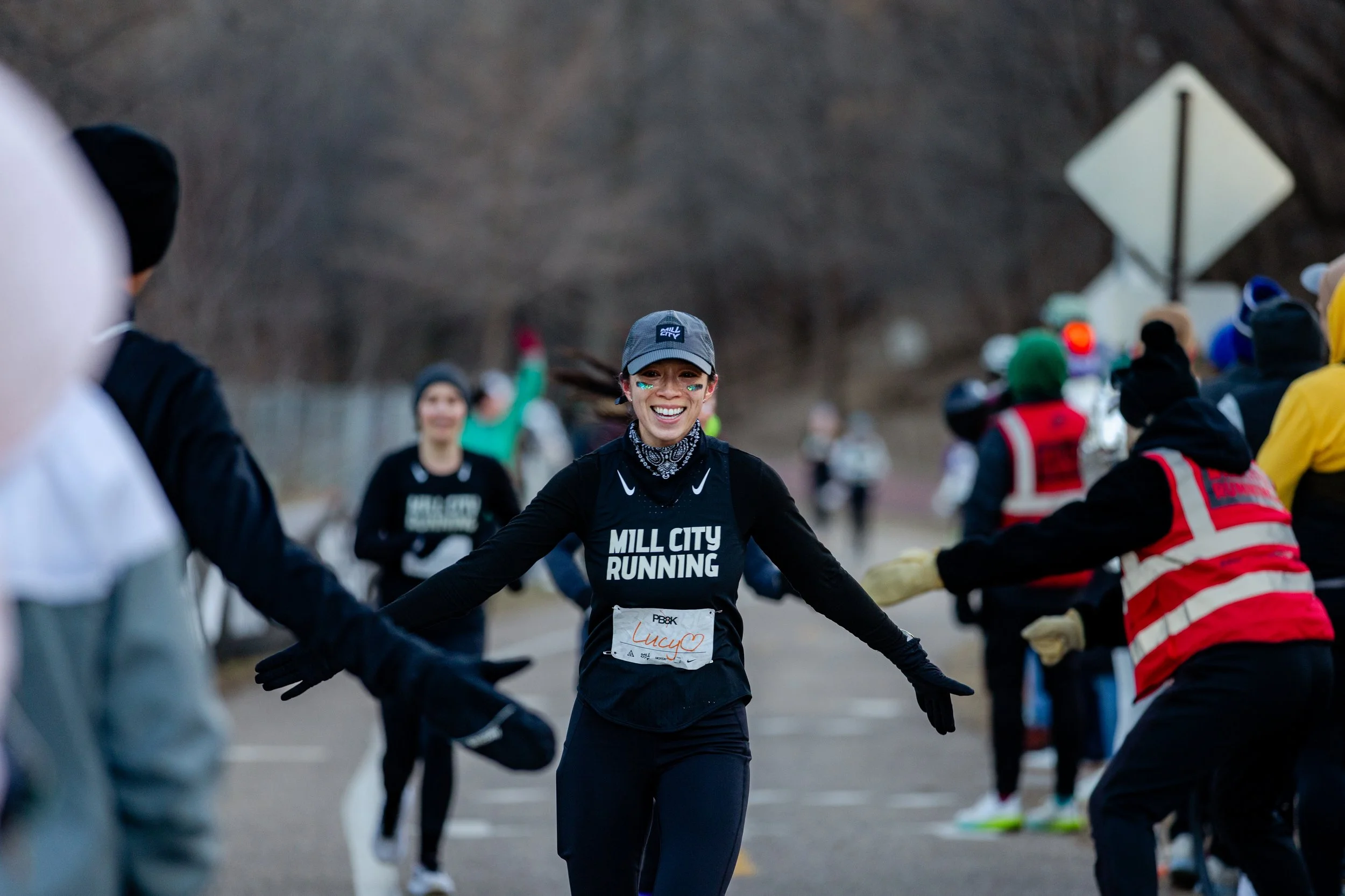 A smiling female runner at a marathon event wearing a black mill city running shirt, gray cap, and black gloves, crossing the finish line with supporters and other runners in the background.