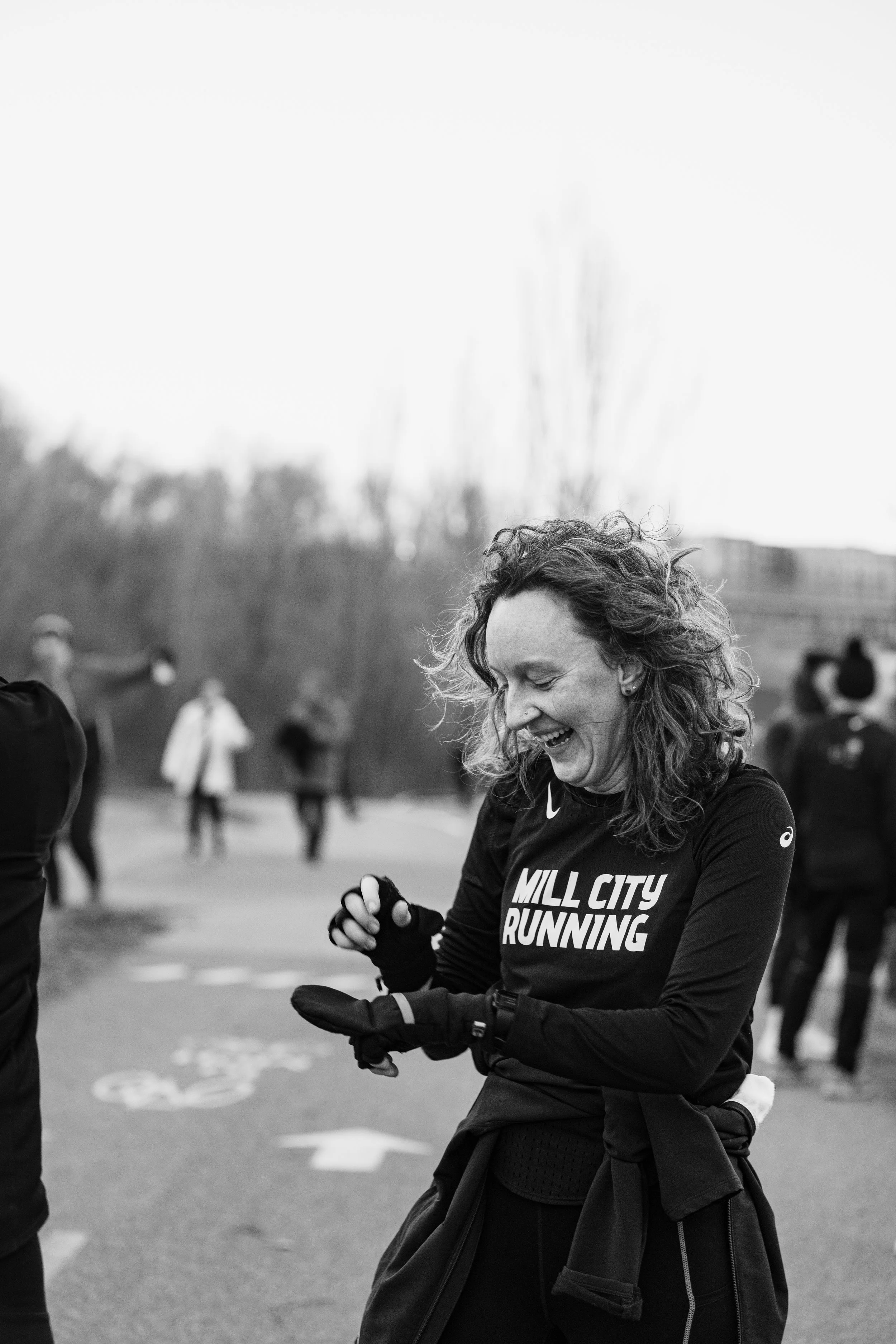 A woman smiling and adjusting her running gloves before a race at Mill City Running, with other runners and trees in the background.