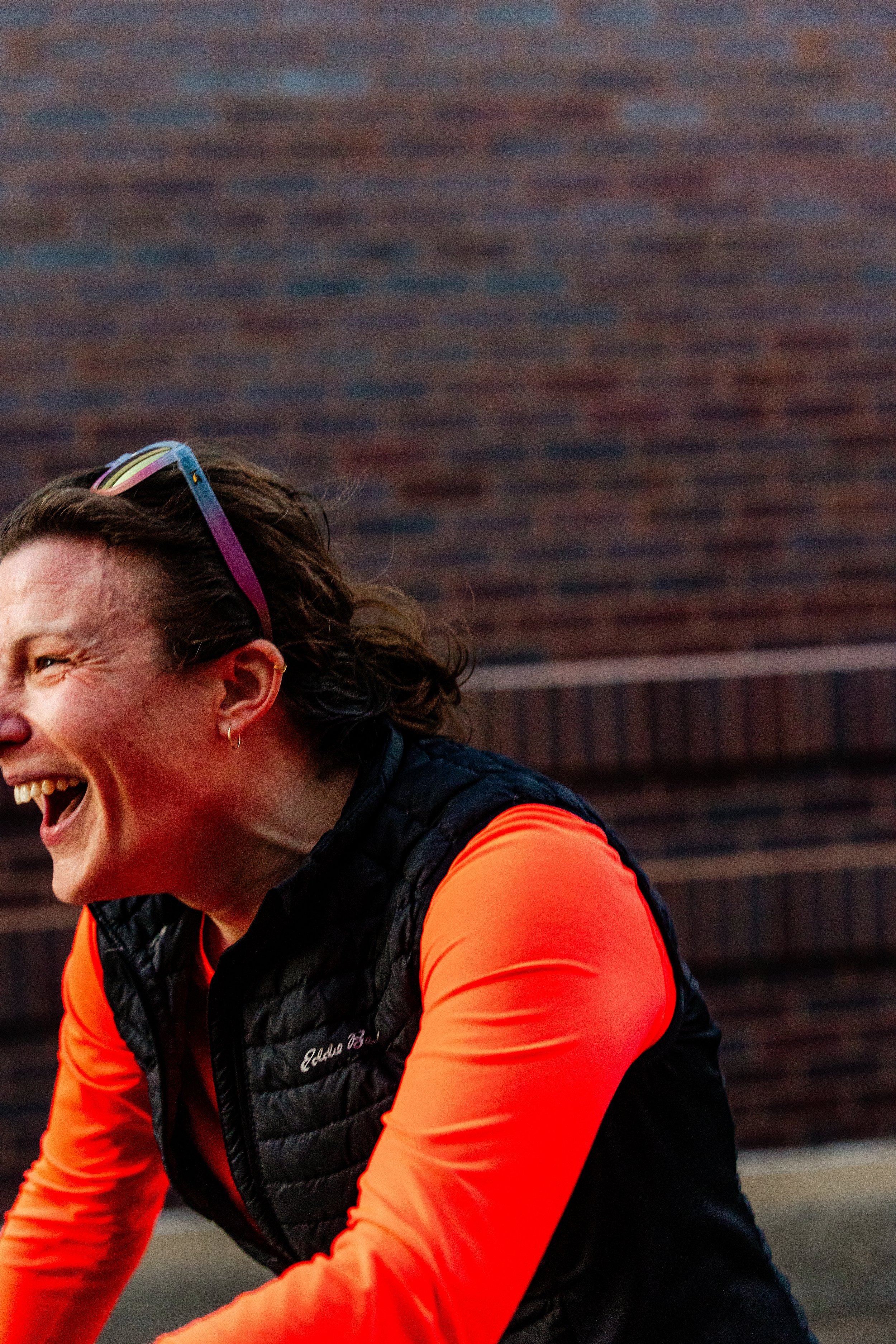 A woman laughing outdoors, wearing an orange long-sleeve shirt, a black vest, and sunglasses on her head, with a blurred brick wall background.