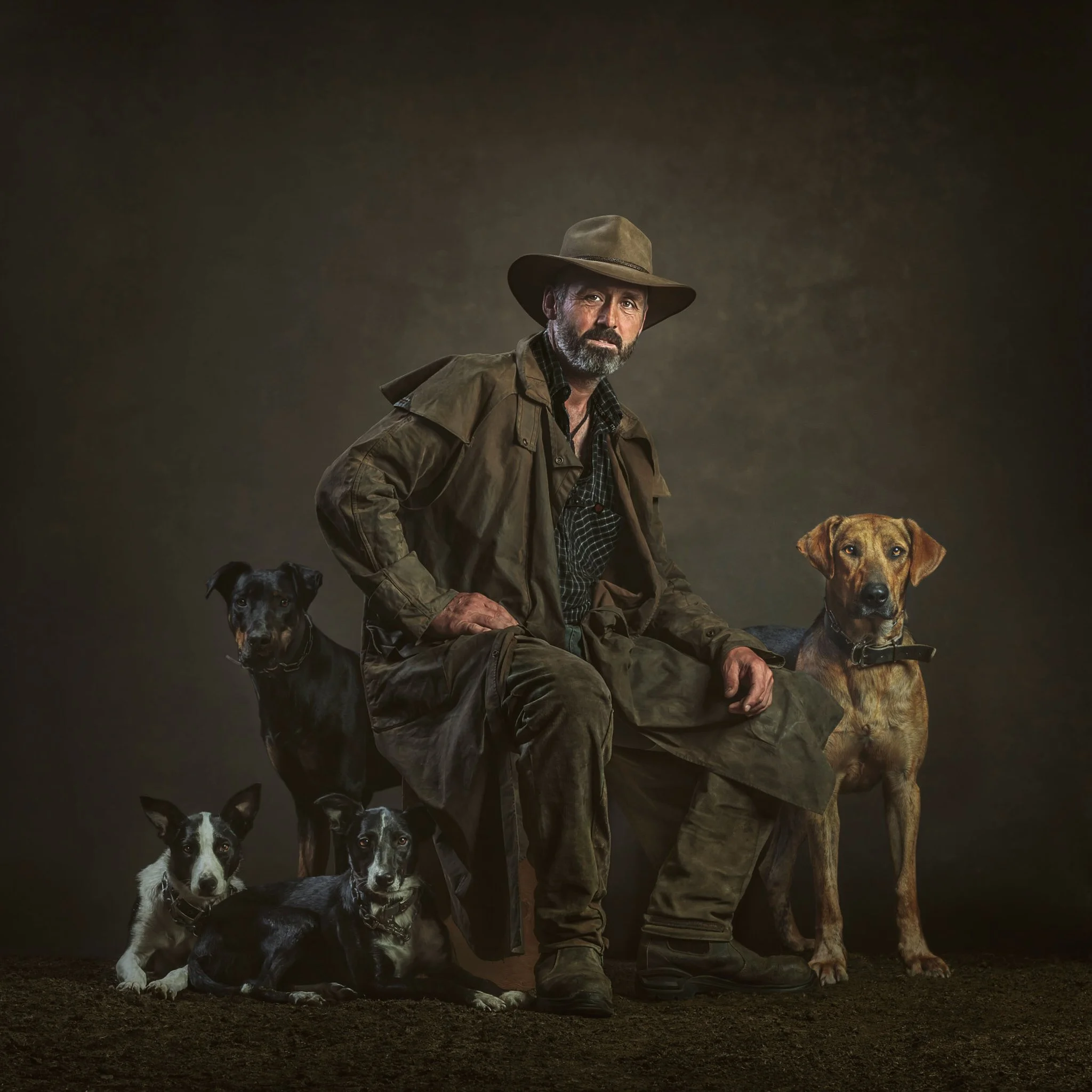 A man dressed in outdoor clothing and a wide-brimmed hat sitting on a stool, surrounded by five dogs, against a dark background.