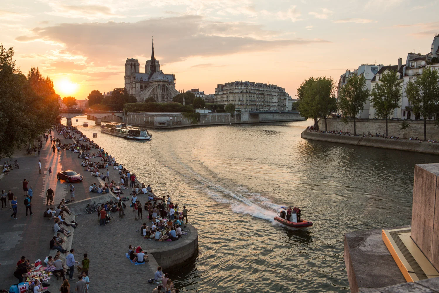 A small speedboat navigates the Seine River in Paris, France. Parisians and tourists line the riverbanks as the sun sets behind the Notre Dame Cathedral.