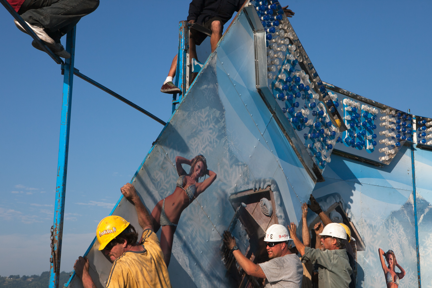 A girl “poses in a bikini” as carnival workers setup a ride called the “Ice Storm.” Teamwork and communication are essential to preclude an accident from happening.