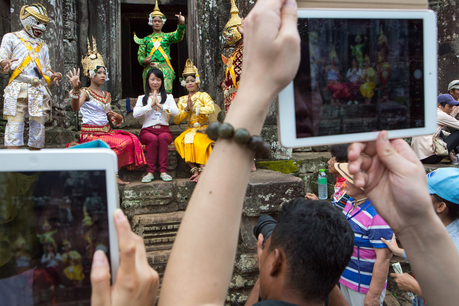 A visitor to the Angkor Thom complex in Cambodia has her picture made with a group of boys and girls who are in traditional costume. Several other visitors wait nearby for their opportunity. The boys and girls dress in costume and make a living by standing in the Angkor complex and posing for pictures with tourists.