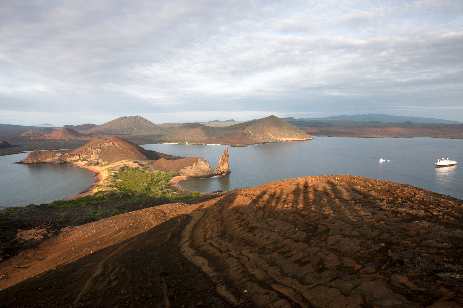 In the Galapagos Islands, the shadows of tourists are visible on the backdrop and landscape of Bartolome Island. An expedition vessels anchors in the water.