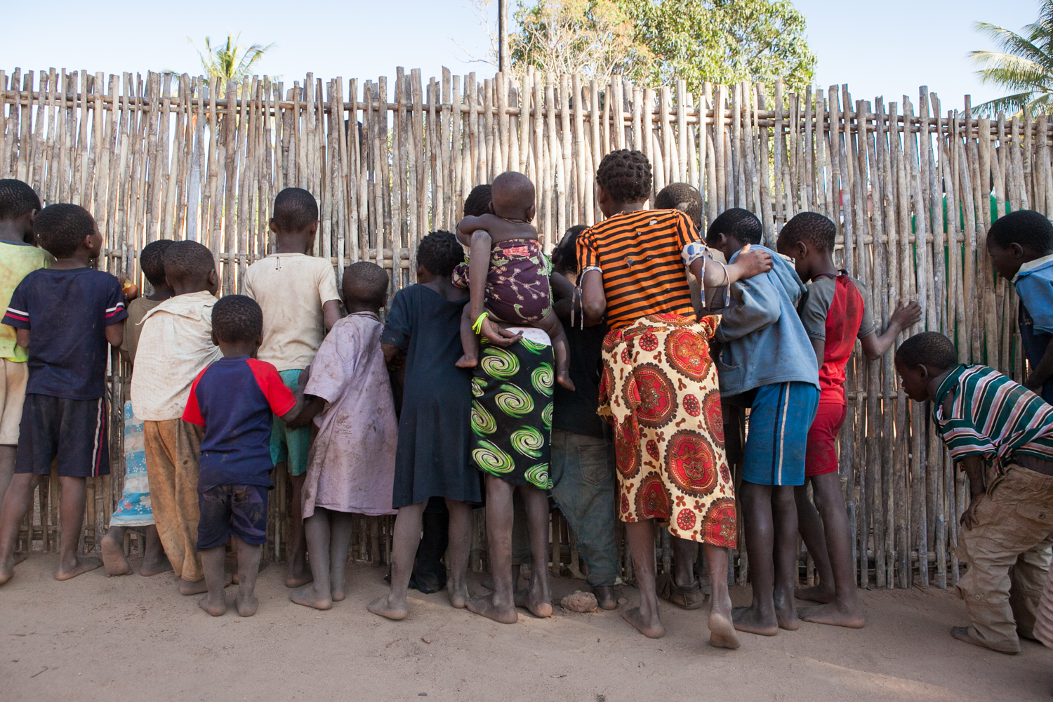  During a pump repair, locals gather around a fence and curiously peer through its cracks. 