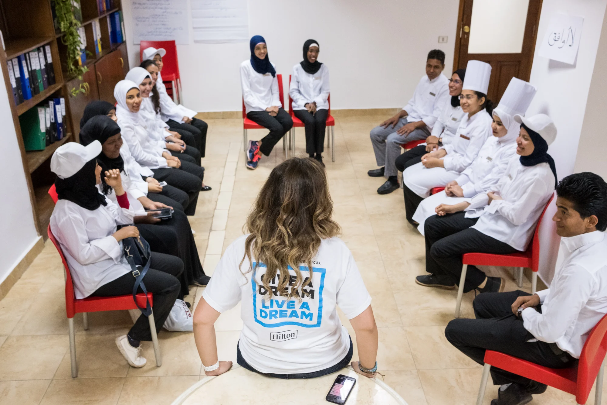 Ingy (leaning on table) speaks with the initial group of 15 participant trainees. Each workday, Ingy has a check-in with the trainees to provide overall direction and receive feedback on their training activities.
