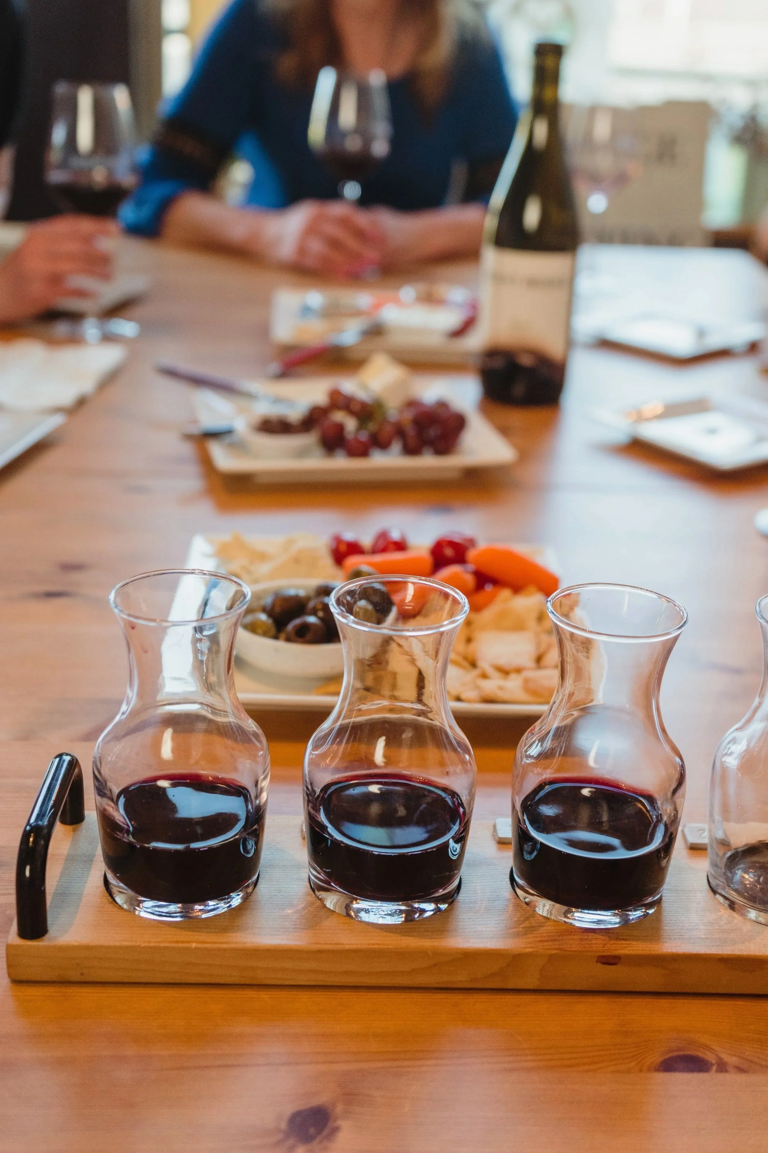 Four glasses of red wine on a wooden serving tray in the foreground, with a tray of cheese, grapes, and vegetables in the background at a gathering.