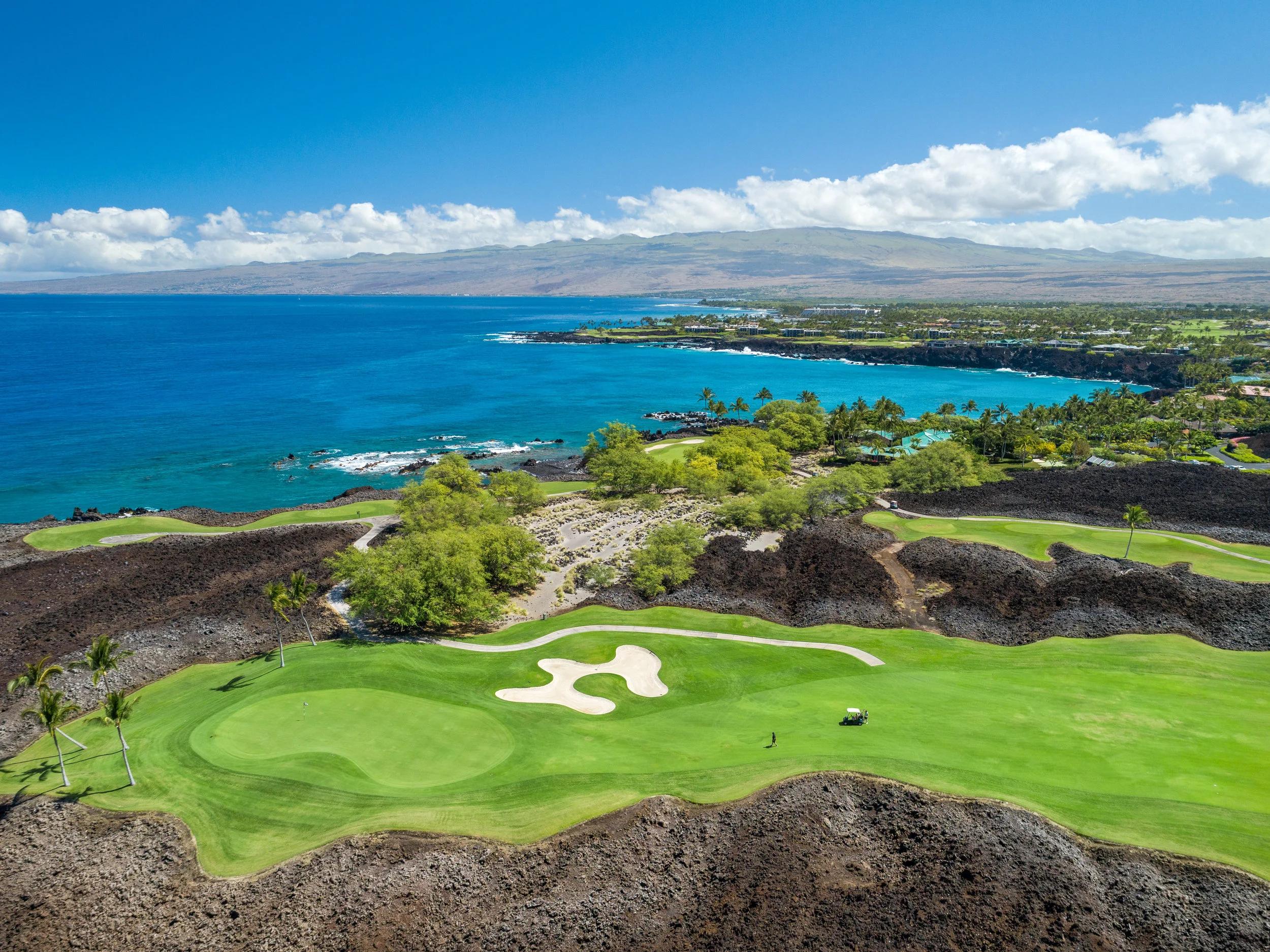 Green fairways against a dramatic black lava backdrop, with ocean and mountain views, as seen from the rear lots at Ke Kailani.