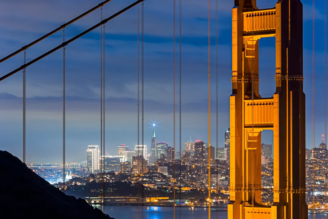 **Christmas Through the Cables**

San Francisco's glowing skyline framed through the Golden Gate Bridge cables at night from Hawk Hill—the city adorned with Christmas lights, creating a festive sparkle through the bridge's iconic orange geometry.

Th