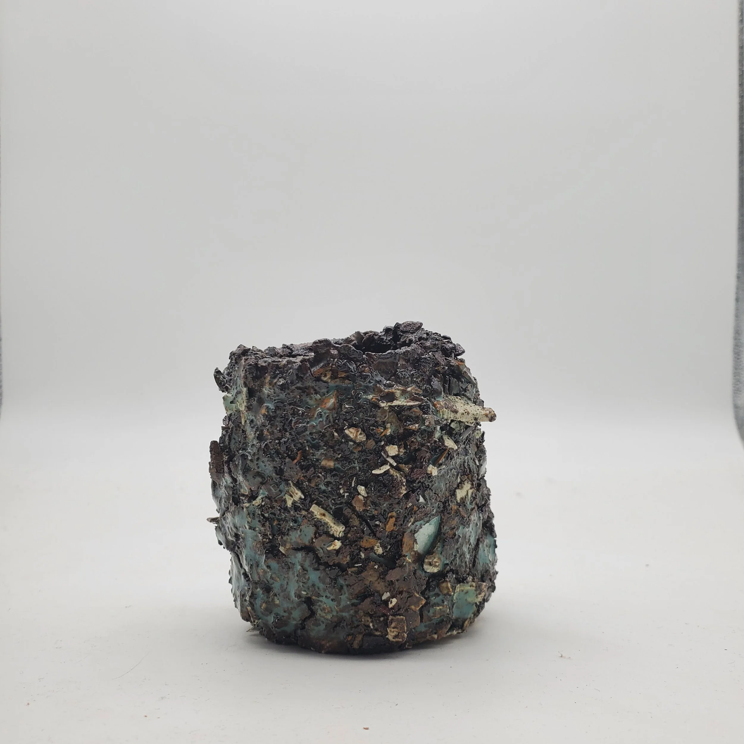 A small, irregularly shaped object with a rough, textured surface featuring various shades of black, brown, and hints of greenish-blue, placed on a plain white background.