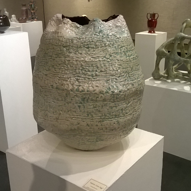 A large, textured ceramic or stone vessel with an uneven, rough surface, displayed on a white pedestal in an art gallery. The vessel has a rounded shape with a wide opening at the top.