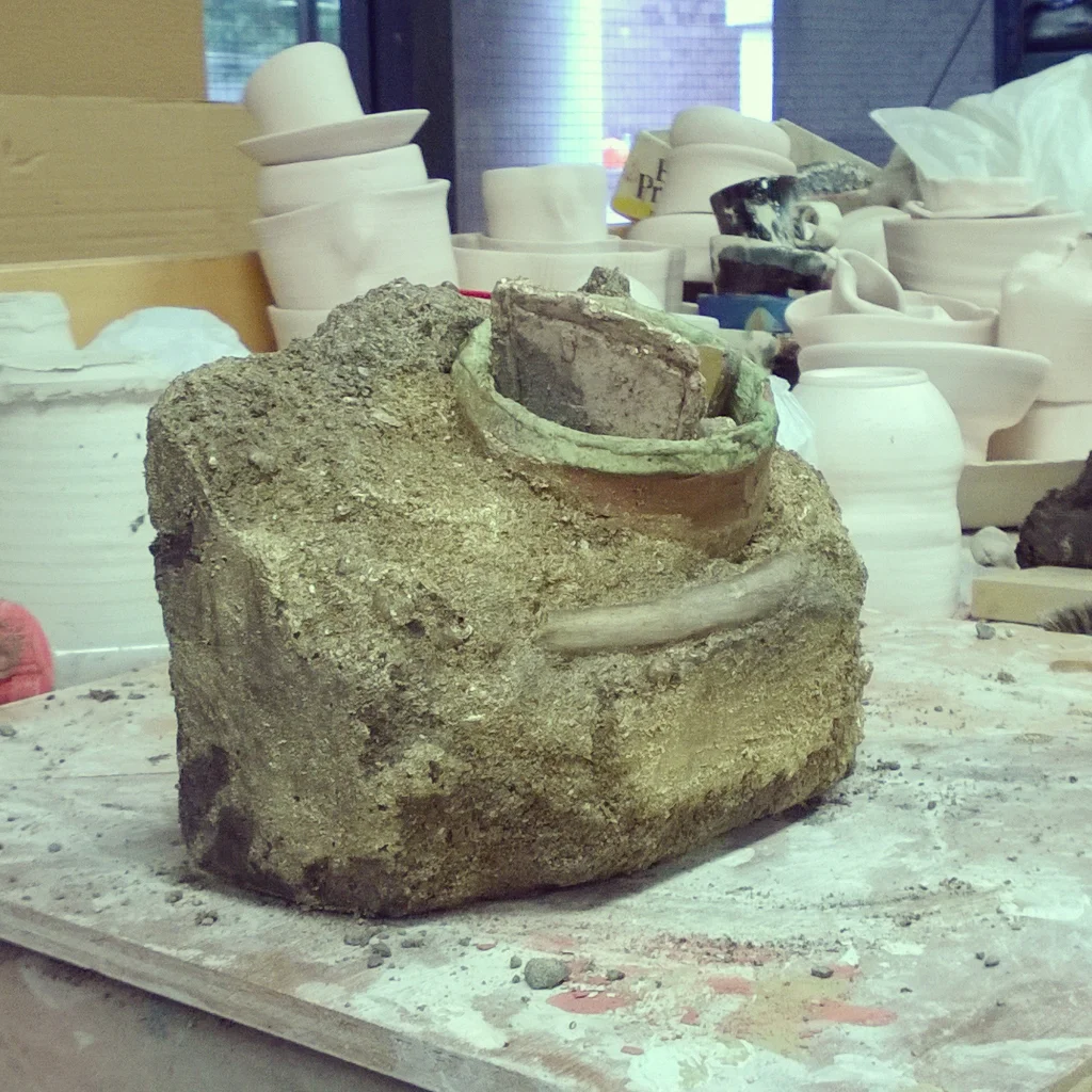 A rough, partially broken ceramic sculpture or object covered in dirt, surrounded by various pottery pieces and molds in a workshop or studio setting.
