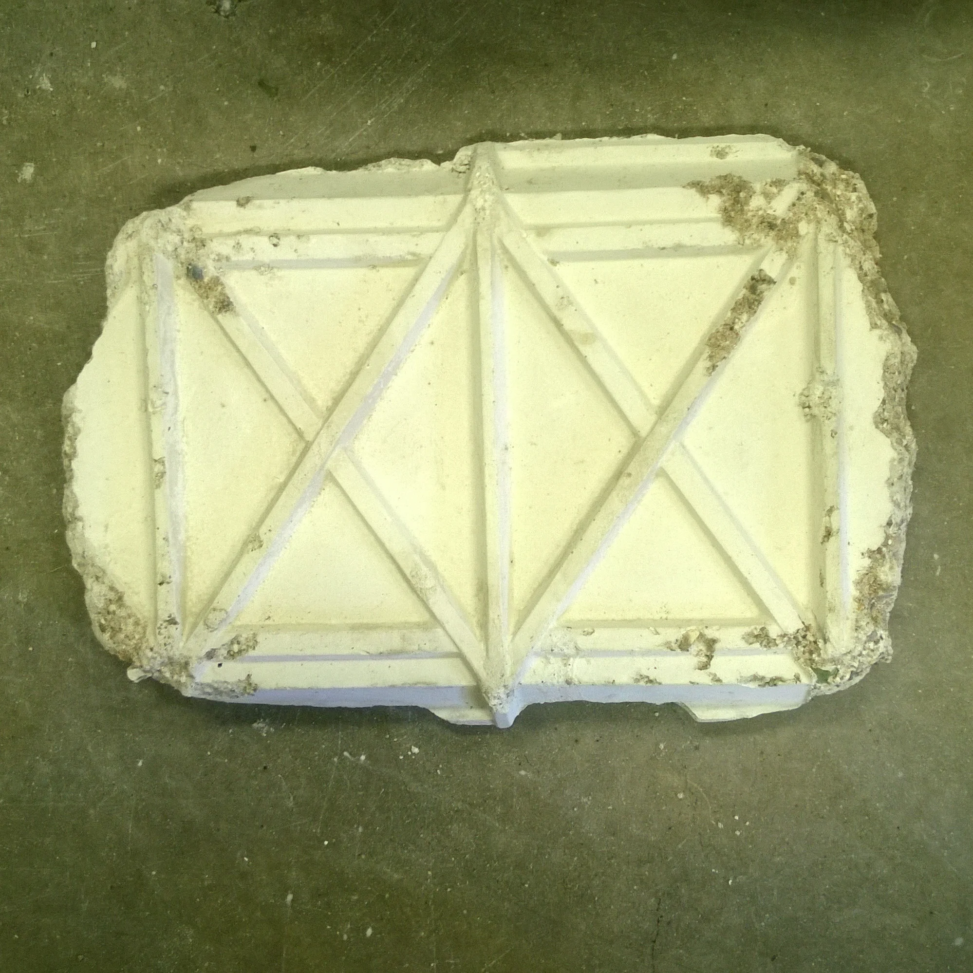 A rectangular piece of drywall with a pattern of raised lines on the surface, partially covered with dirt and debris, placed on a concrete floor.