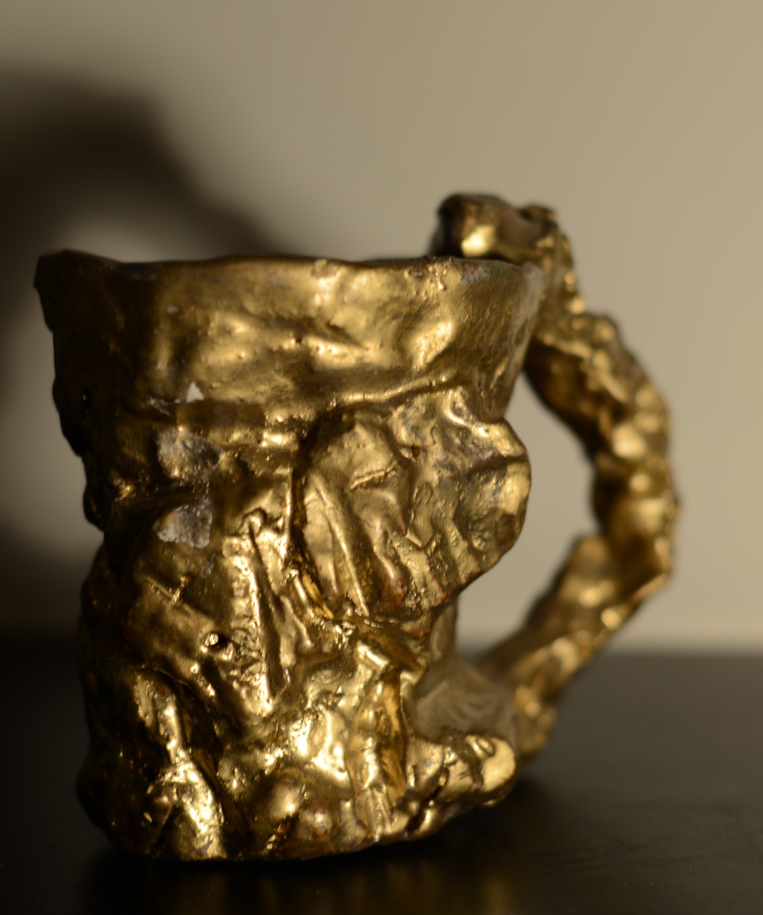 Close-up photo of a gold-colored, textured cup with an irregular surface, placed on a dark surface against a plain background.