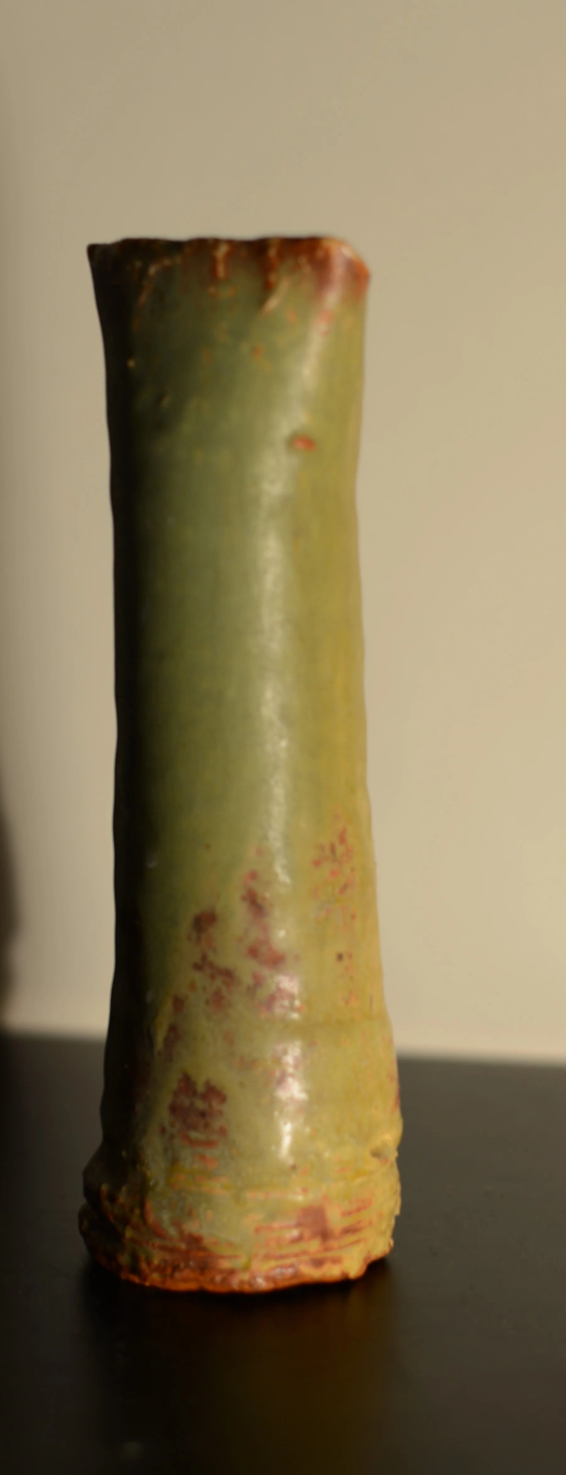 Close-up of a green and brown glazed ceramic vase with a textured surface, placed on a black surface against a plain background.