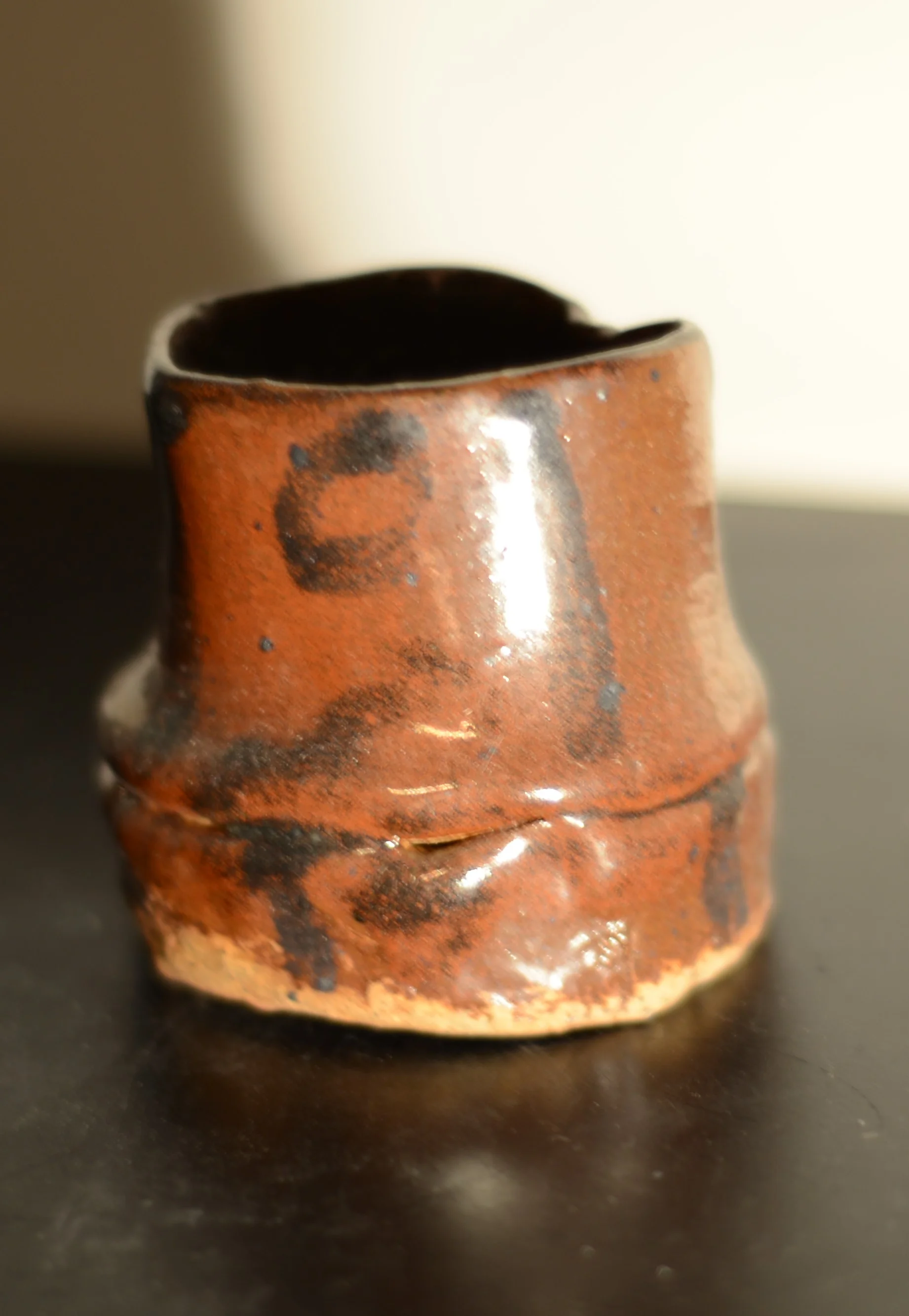 Close-up image of a small, weathered, reddish-brown ceramic cup with black spots, sitting on a dark surface.