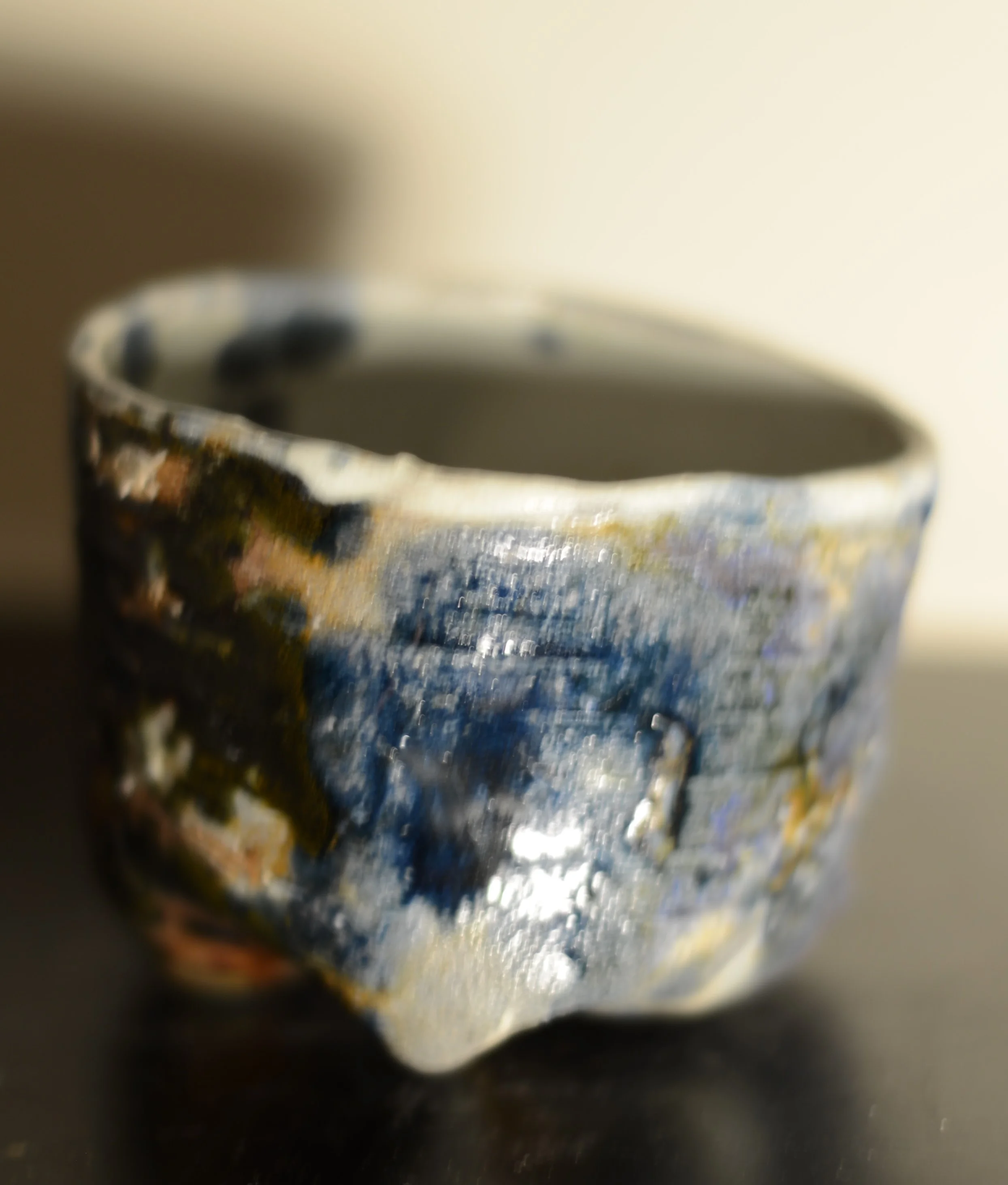 Close-up of a small ceramic bowl with a textured, multicolored glaze in shades of blue, white, and earthy tones, placed on a reflective black surface.