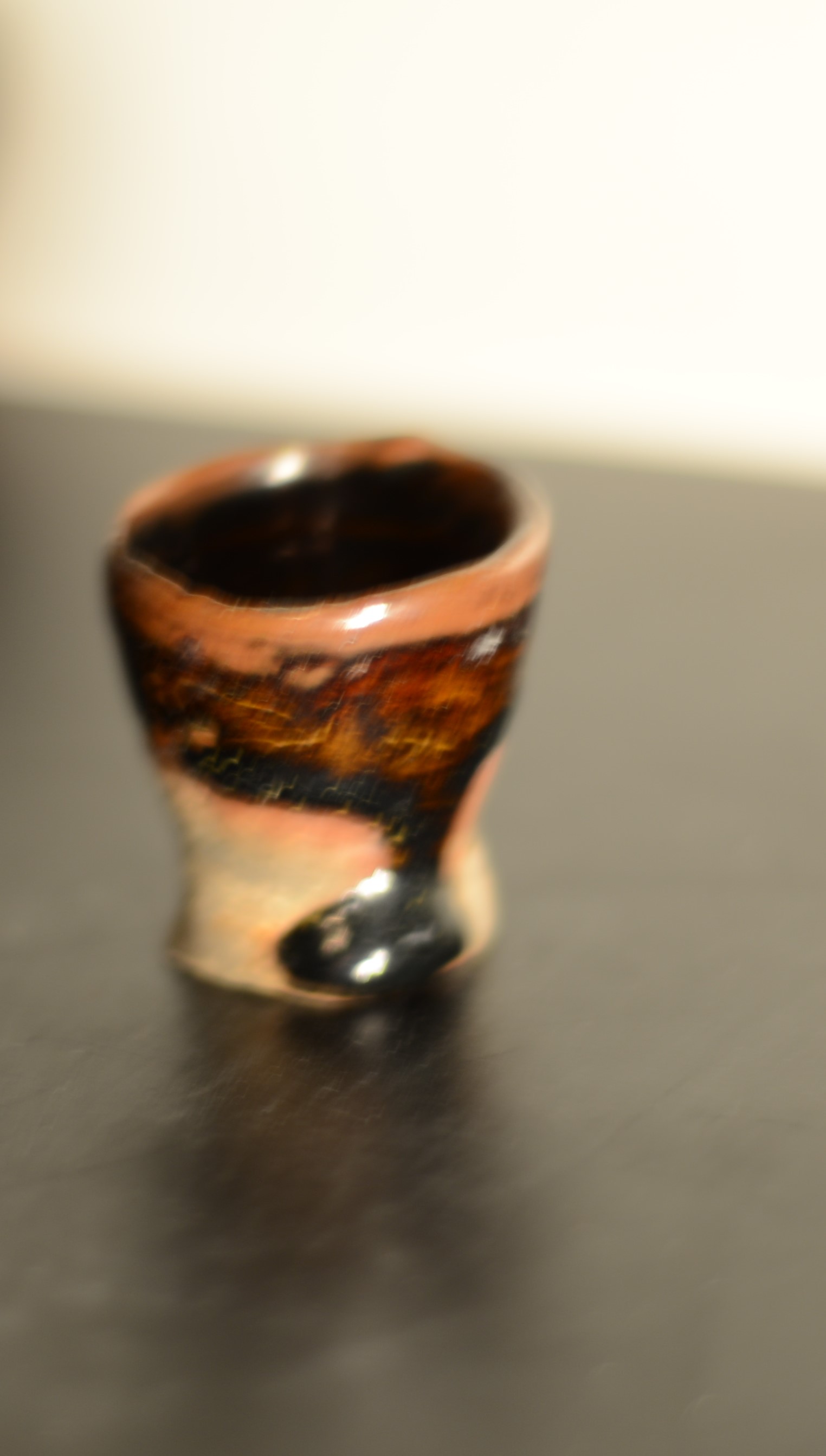 A small, handmade ceramic cup with a glossy finish, featuring a dark brown and black speckled pattern, placed on a dark surface against a blurred background.
