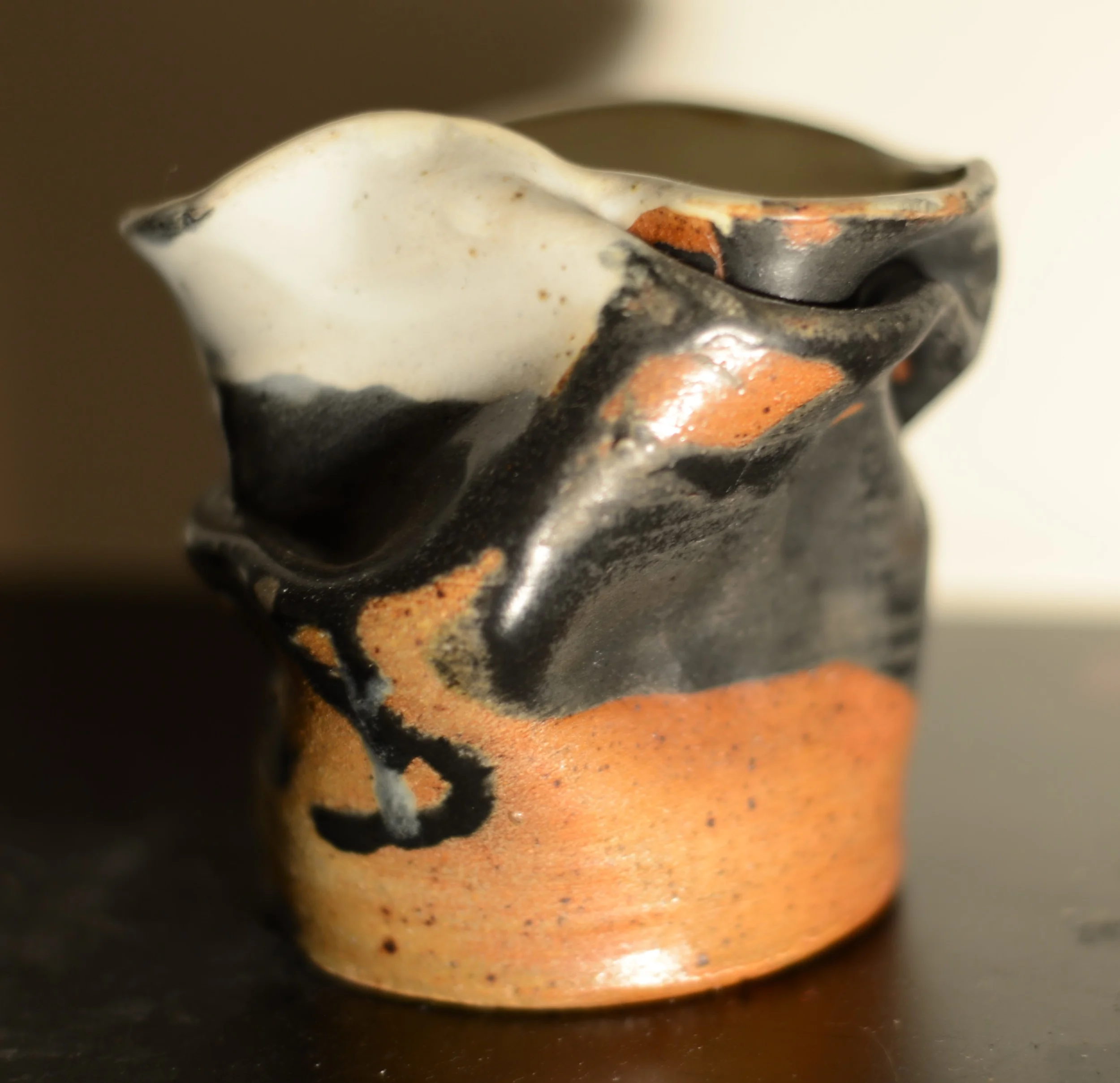 Close-up of a small ceramic pitcher with a v-shaped spout, decorated in black, white, and orange-glazed patterns.
