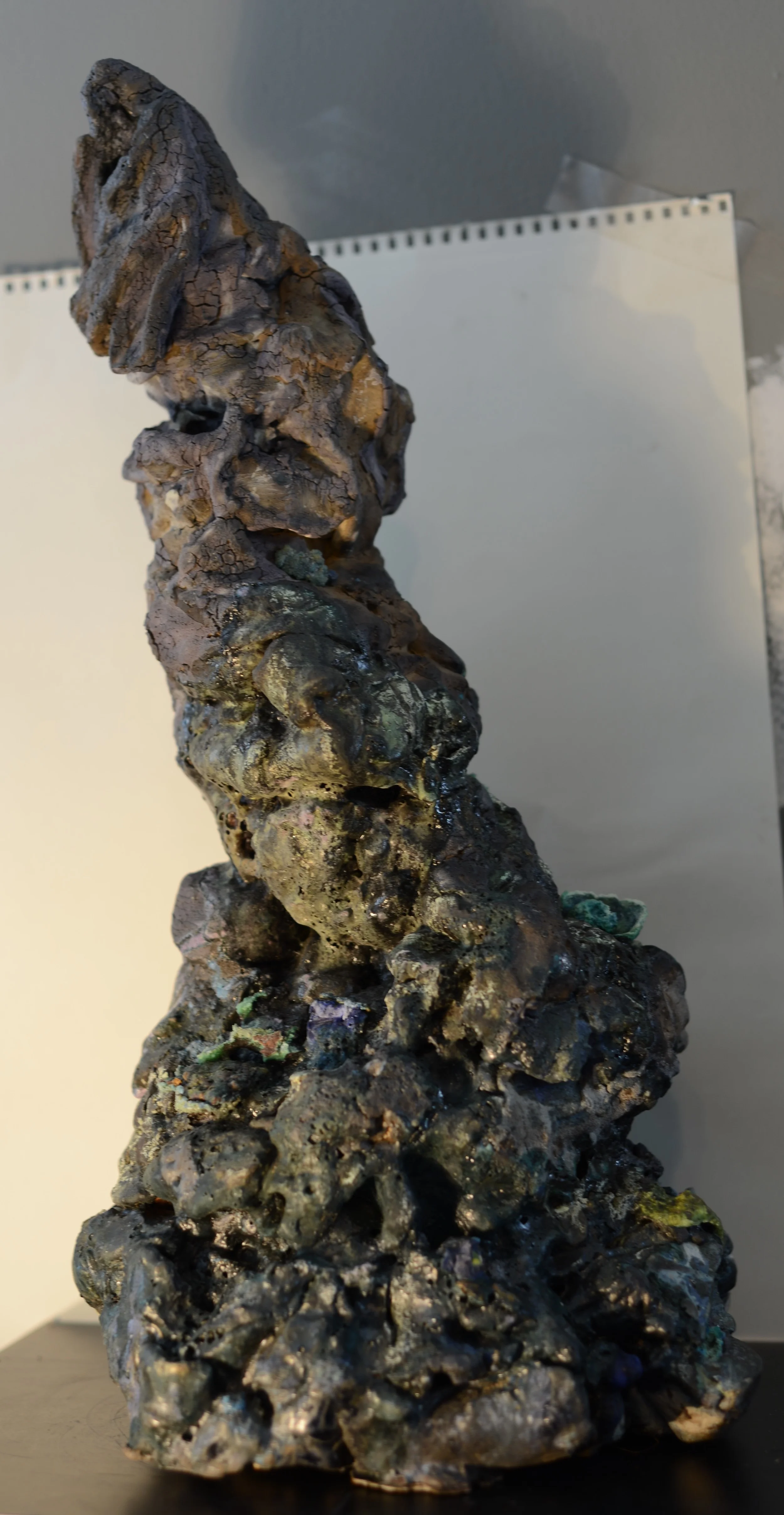 A tall, irregularly shaped rock or mineral formation with a dark, textured surface and patches of shiny, metallic elements, displayed against a plain background.