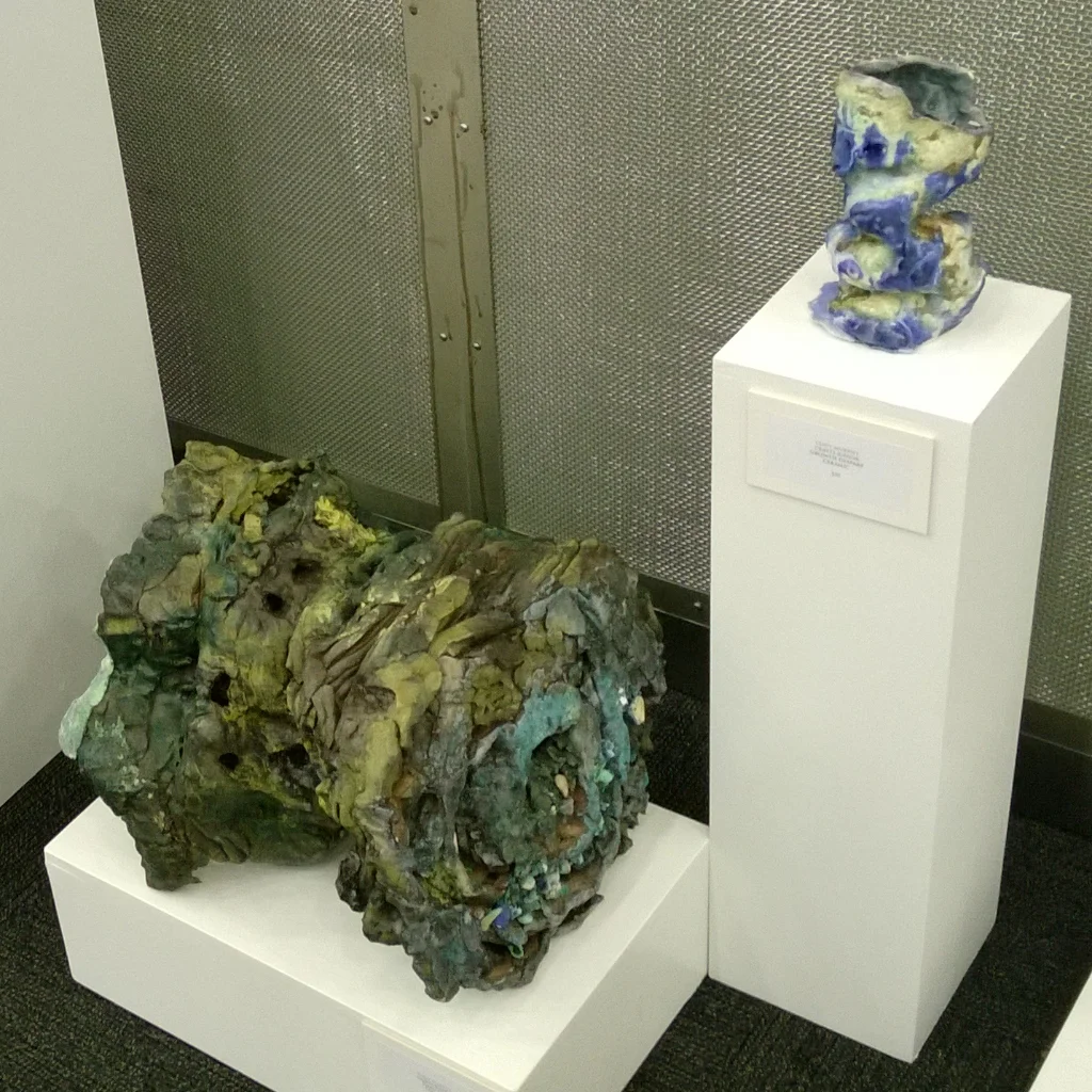 Two abstract, ceramic sculptures on white pedestals, one larger and more irregular, the other smaller with a blue and yellow glaze, in an art gallery setting.