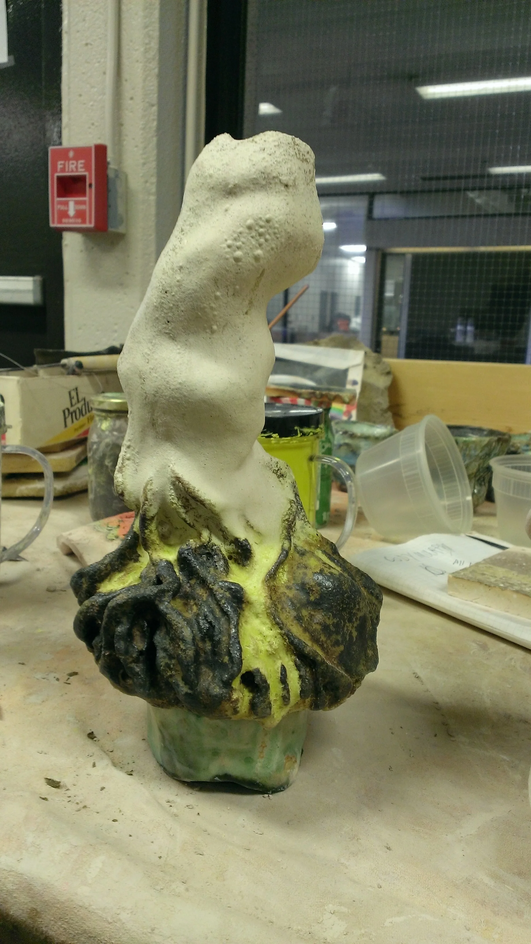 A ceramic or sculpture piece with a rough, textured surface and a painted base, resembling a human torso or abstract figure, on a worktable with art supplies in the background.