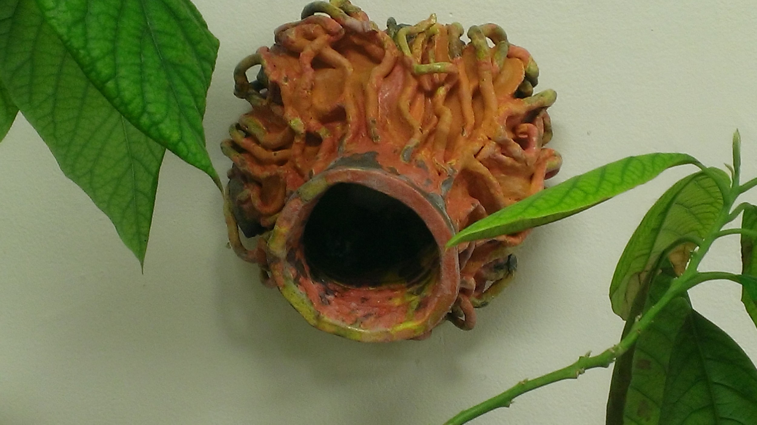 A decorative ceramic birdhouse with a textured, curly orange and green surface, mounted on a wall surrounded by green leaves.