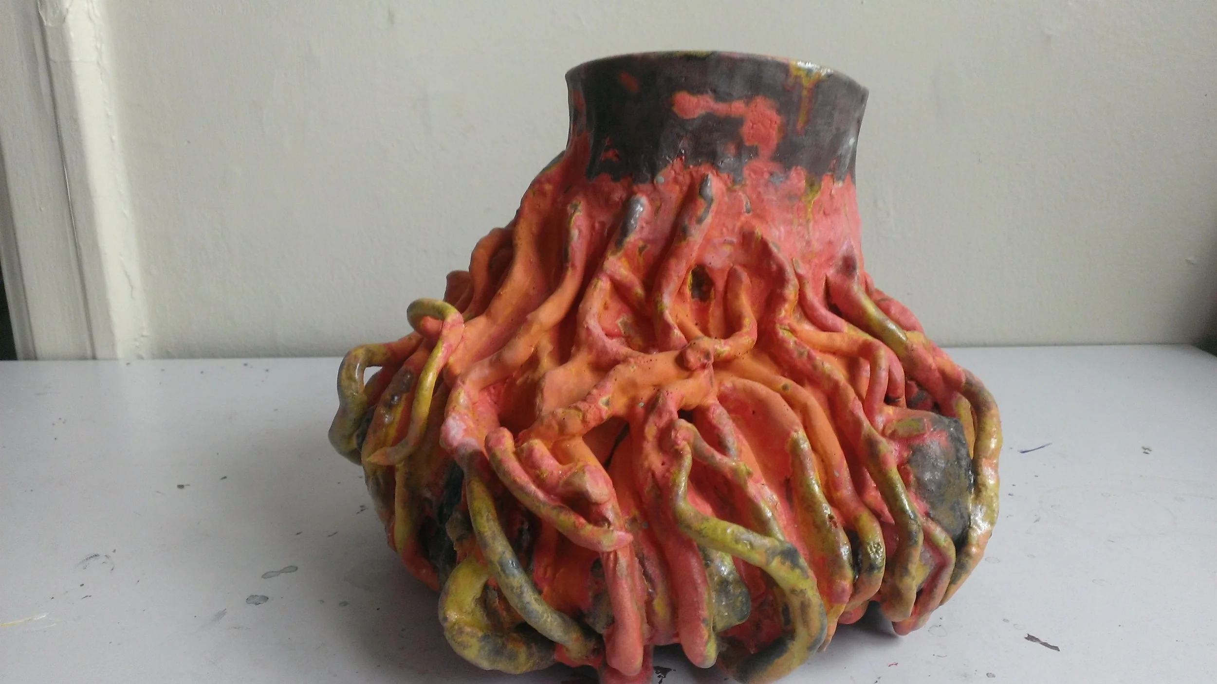 Artistic ceramic vase with a textured surface resembling tree roots, painted in shades of pink, orange, yellow, and black, sitting on a white surface against a plain wall.
