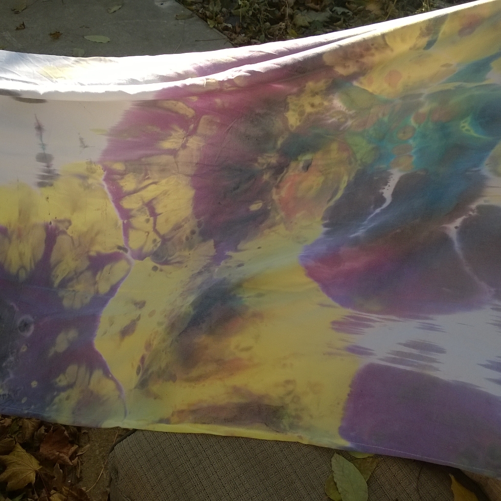 Close-up of a colorful tie-dye fabric with purple, yellow, green, and blue hues, draped outdoors on fallen leaves.