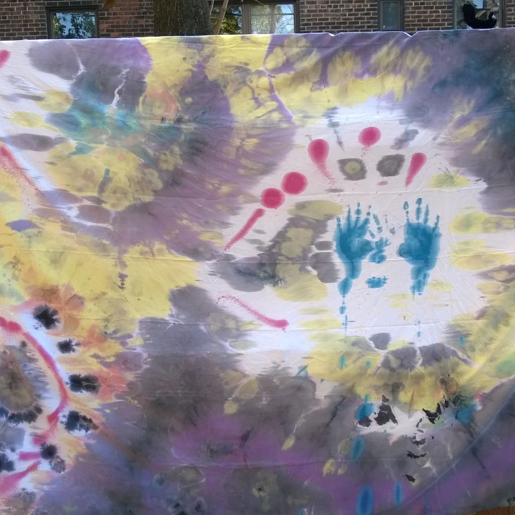 Colorful tie-dye fabric featuring splashes of yellow, purple, blue, red, black, gray, and green with handprints and abstract shapes.