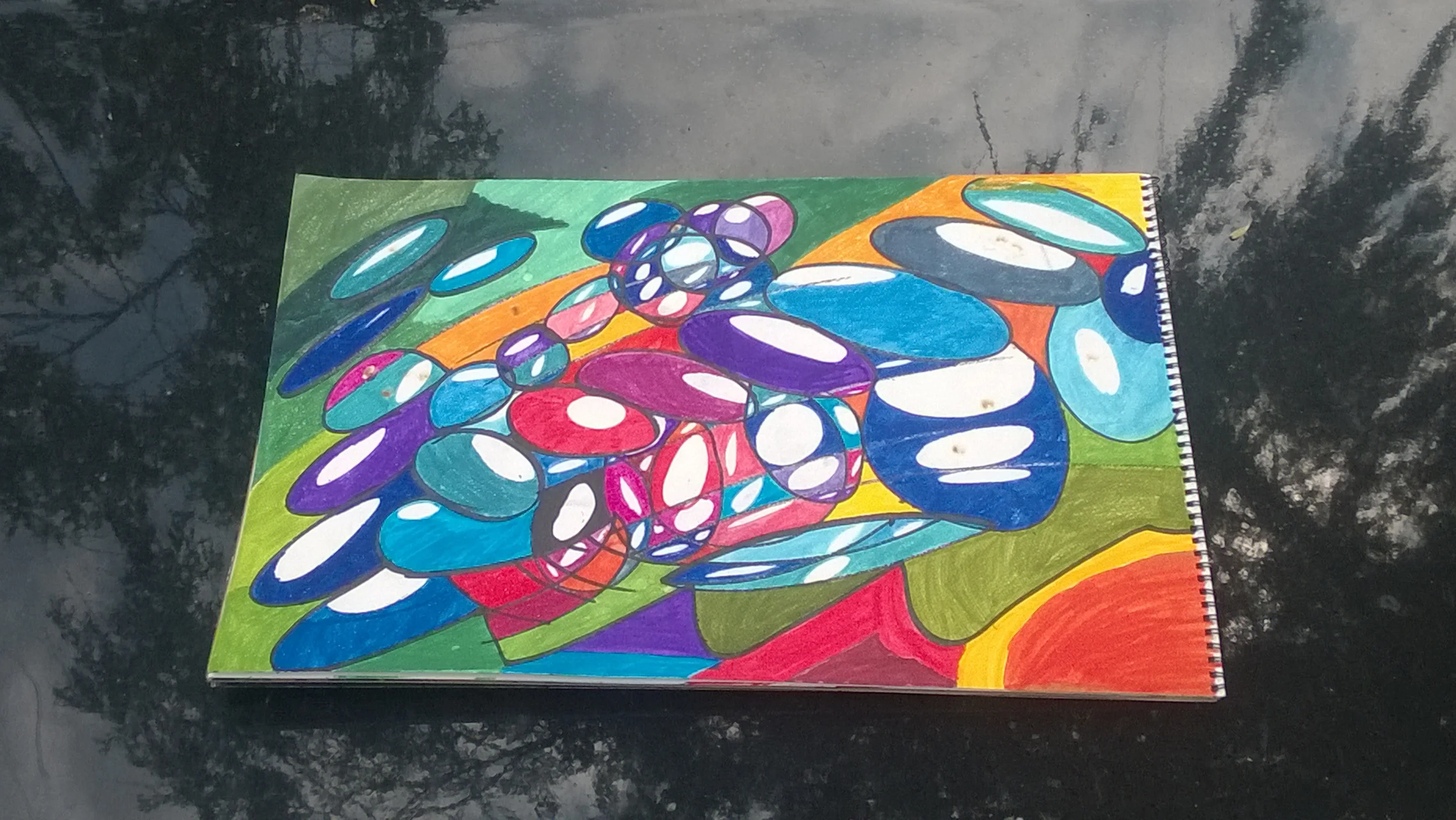 Colorful abstract drawing with oval shapes in purple, blue, pink, red, white, and black on paper, placed on a dark reflective surface. The drawing includes various overlapping ovals and bright background sections.