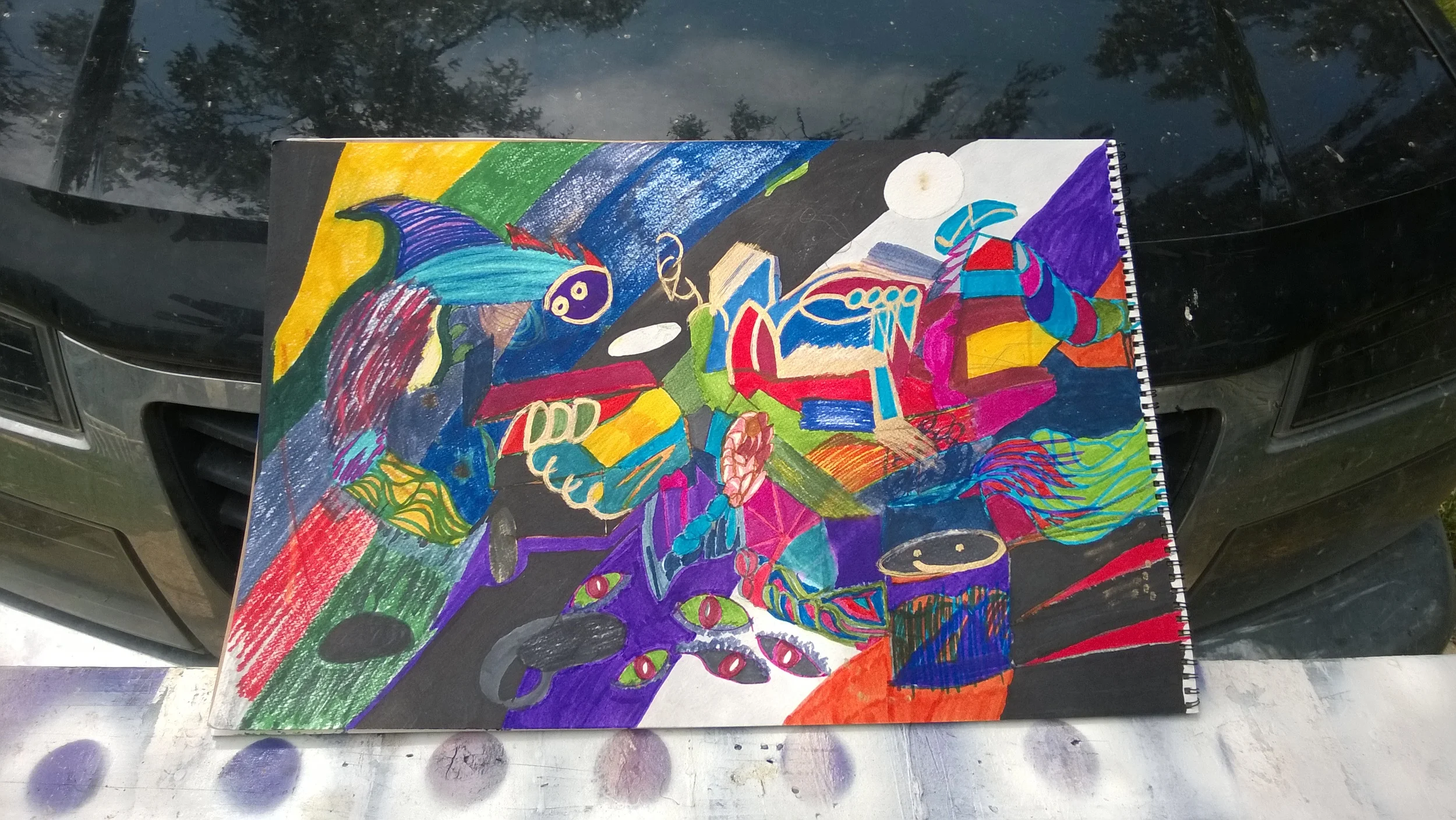A colorful abstract drawing on a notebook placed on a car hood, featuring various shapes, patterns, and vivid colors like red, blue, purple, yellow, and green.