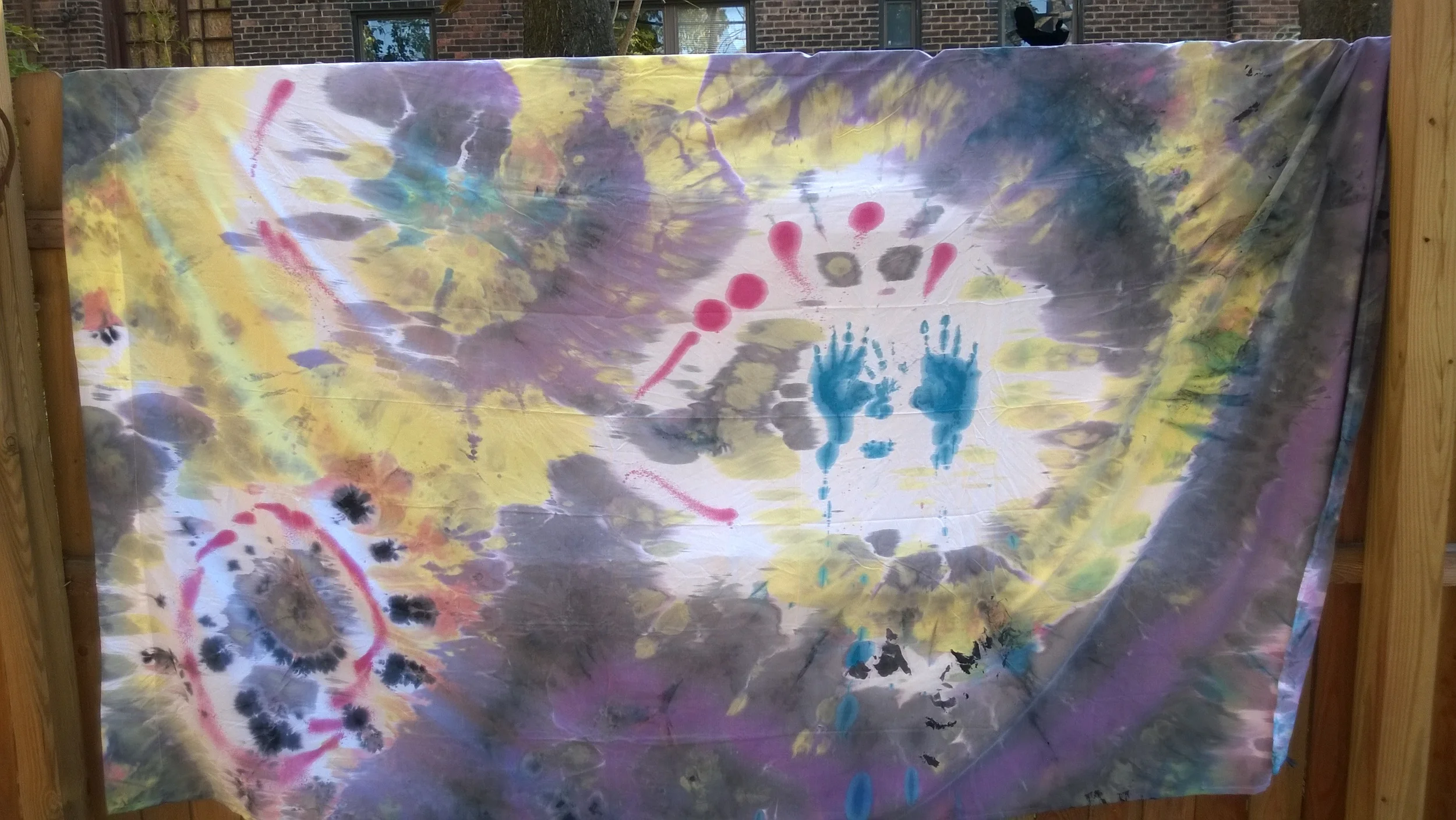 A tie-dye fabric with colorful splashes of yellow, purple, blue, pink, black, and white, hanging on a wooden frame outdoors.