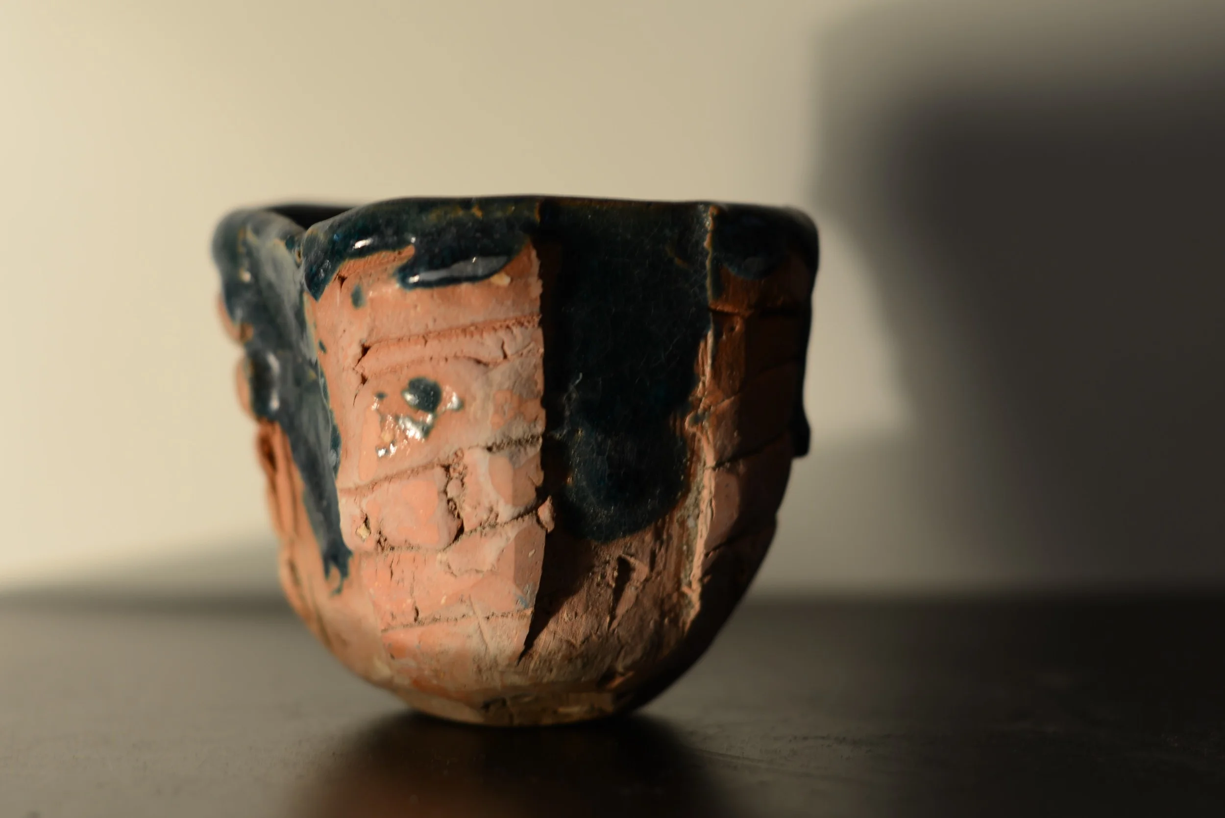 A small ceramic bowl with a rough, textured surface. The bowl is decorated with black and pinkish-orange glaze, featuring a face design with black eyes, nose, and mouth.