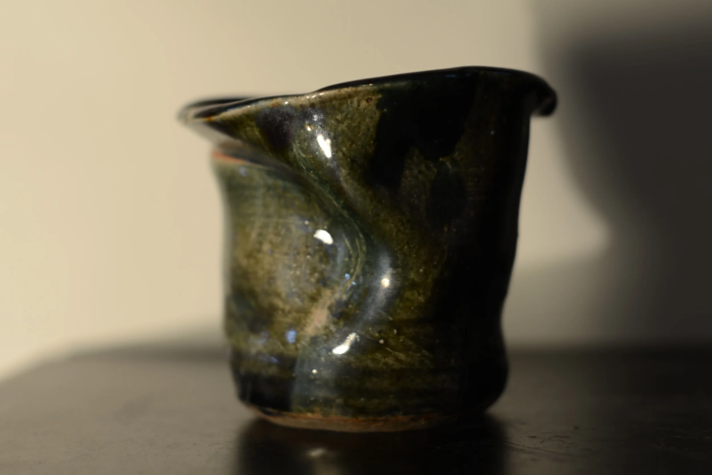 A close-up of a dark, glossy, handmade ceramic vessel with an uneven surface and irregular shape, placed on a dark surface with a plain, light-colored wall in the background.