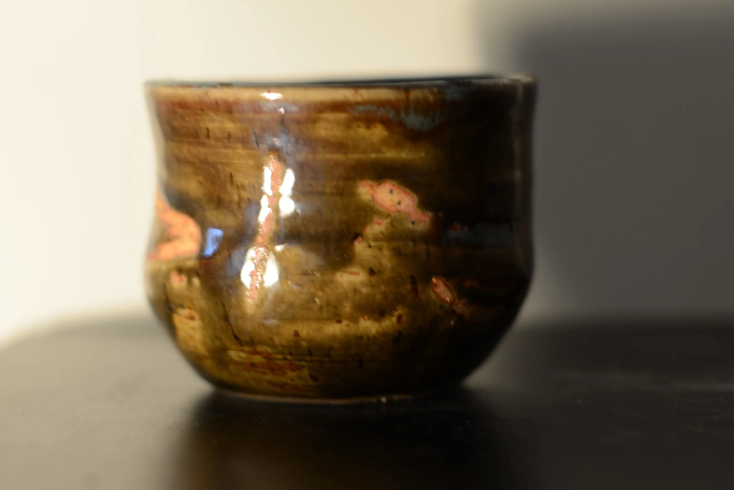 A small, round ceramic bowl with a glossy, glazed finish in shades of brown with pinkish accents, placed on a black surface against a plain light-colored background.