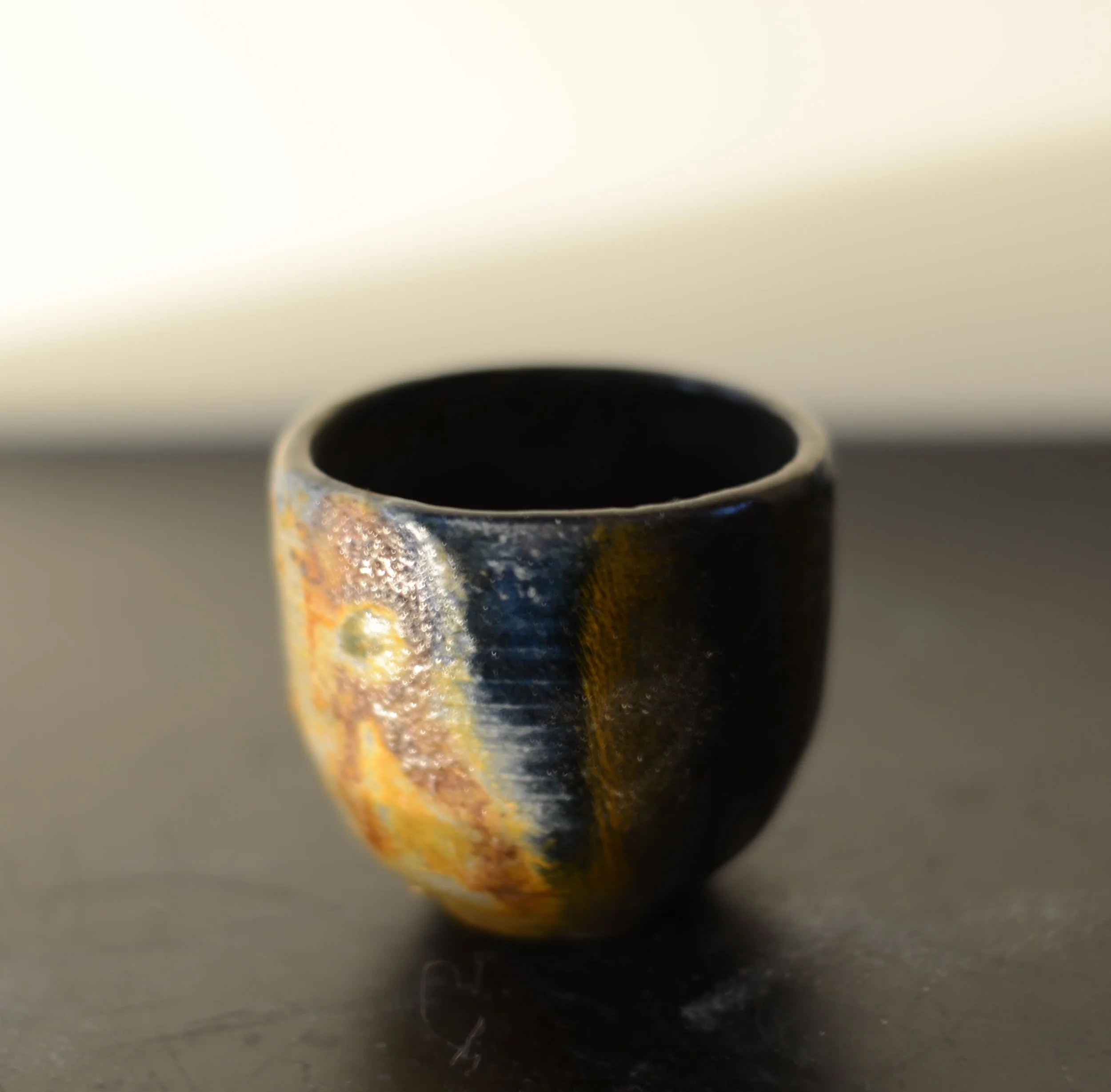 A small ceramic cup with a textured, multicolored glaze on a dark surface.