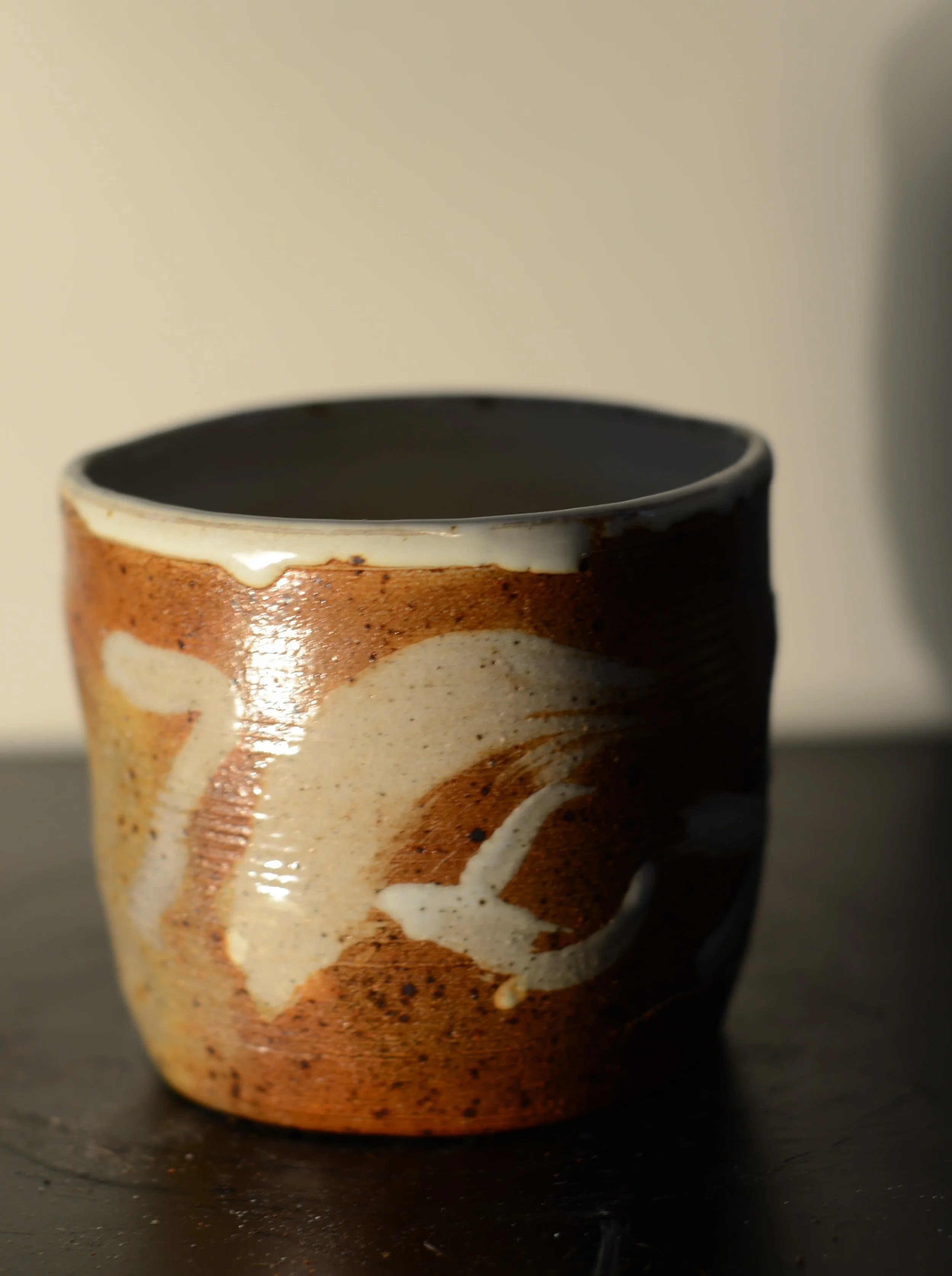 A small ceramic mug with a brown and beige glaze, featuring abstract white swirl pattern, sitting on a dark surface with a light background.