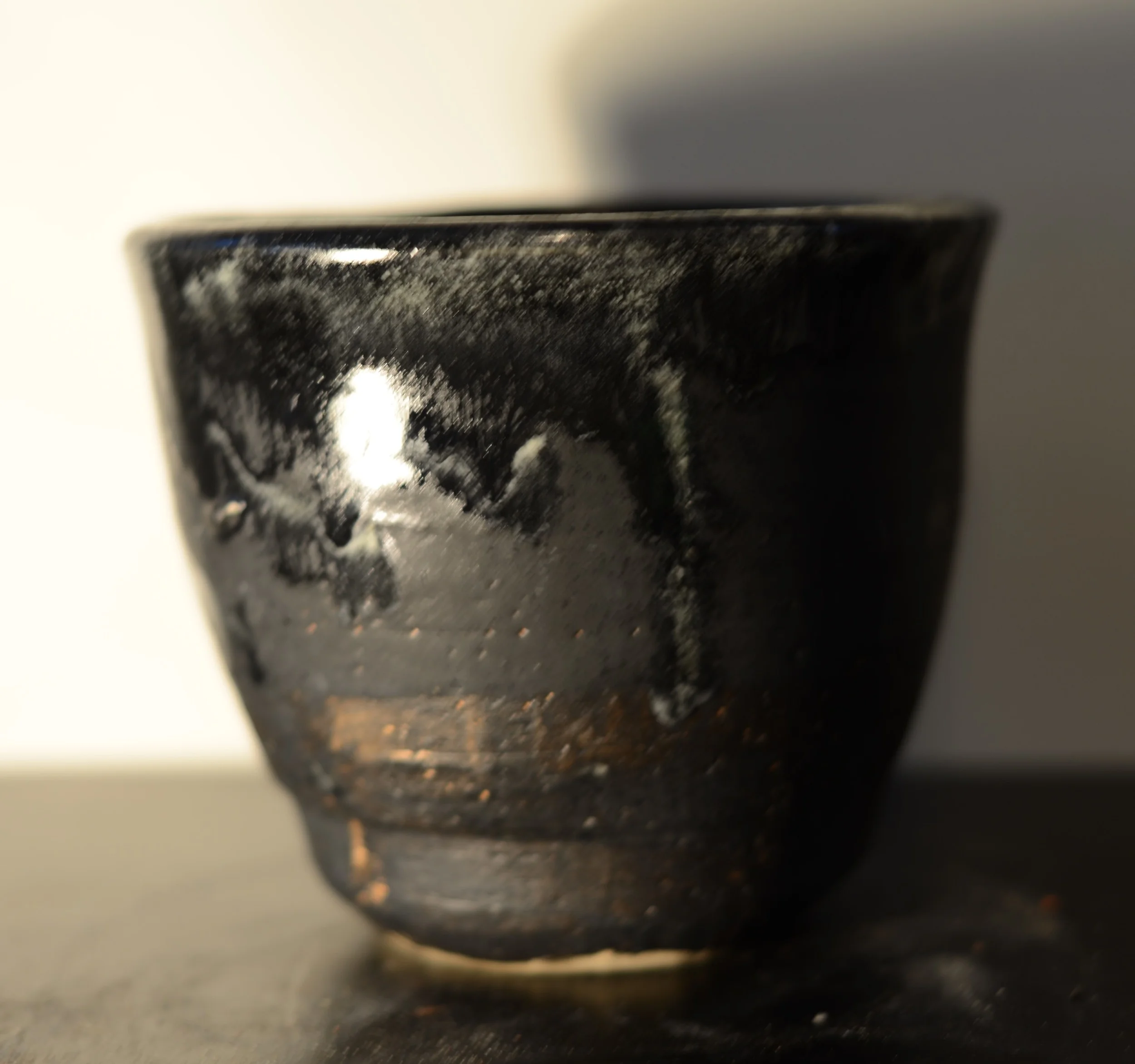 Close-up of a black, ceramic or clay cup with a slightly rough surface and some white and brown markings, placed on a flat dark surface against a light background.