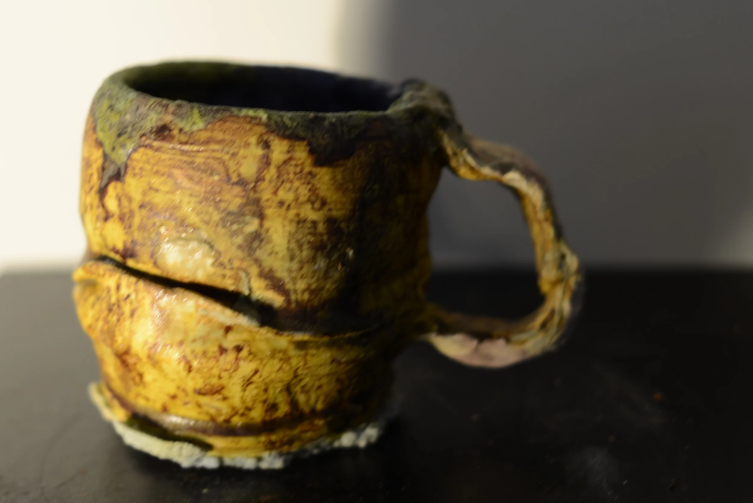 Close-up of a ceramic mug with an uneven, textured surface and a twisted handle, featuring a yellow, brown, and black glaze. The mug is placed on a dark surface against a neutral background.